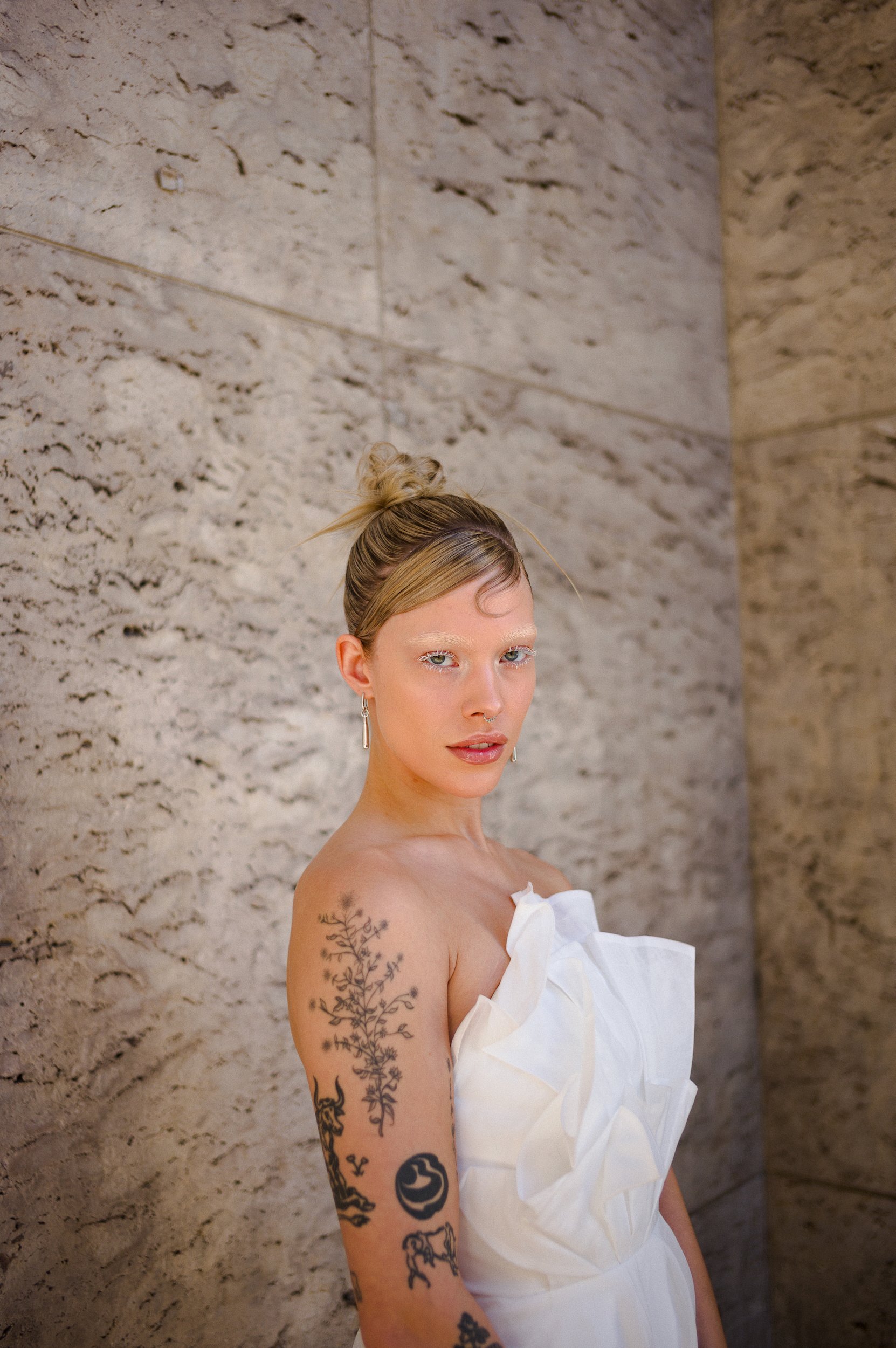 At Paris Fashion Week, A woman with blonde hair styled in a messy bun, wearing a white off-shoulder dress with ruffles, standing against a textured beige wall. She has tattoos on her left arm and earring in her right ear.