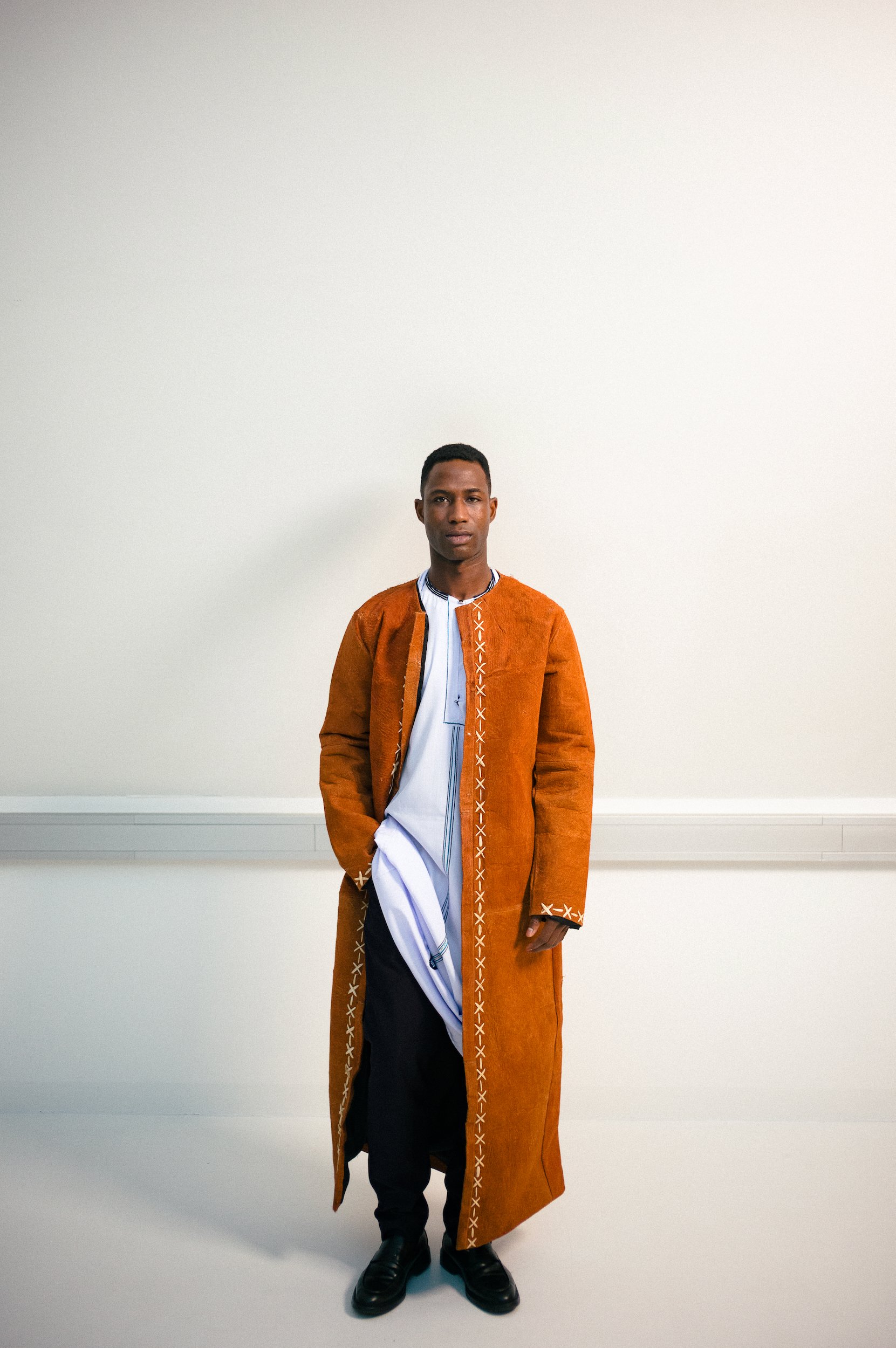 A young man standing against a plain white wall, wearing a long orange coat with decorative stitching, black pants, black shoes, and a white and blue traditional or formal shirt underneath.