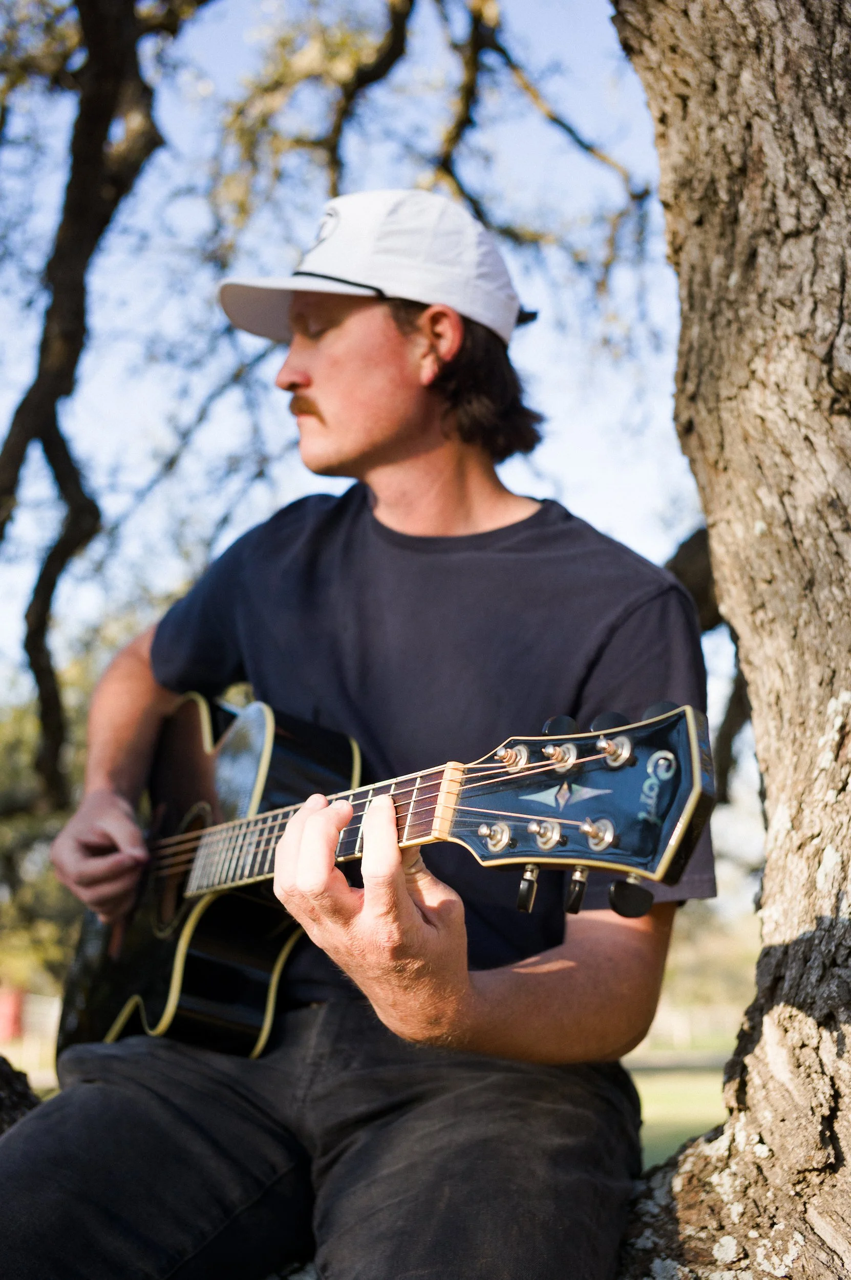 A man sitting outdoors near a tree, playing an acoustic guitar, wearing a white cap and a black t-shirt.