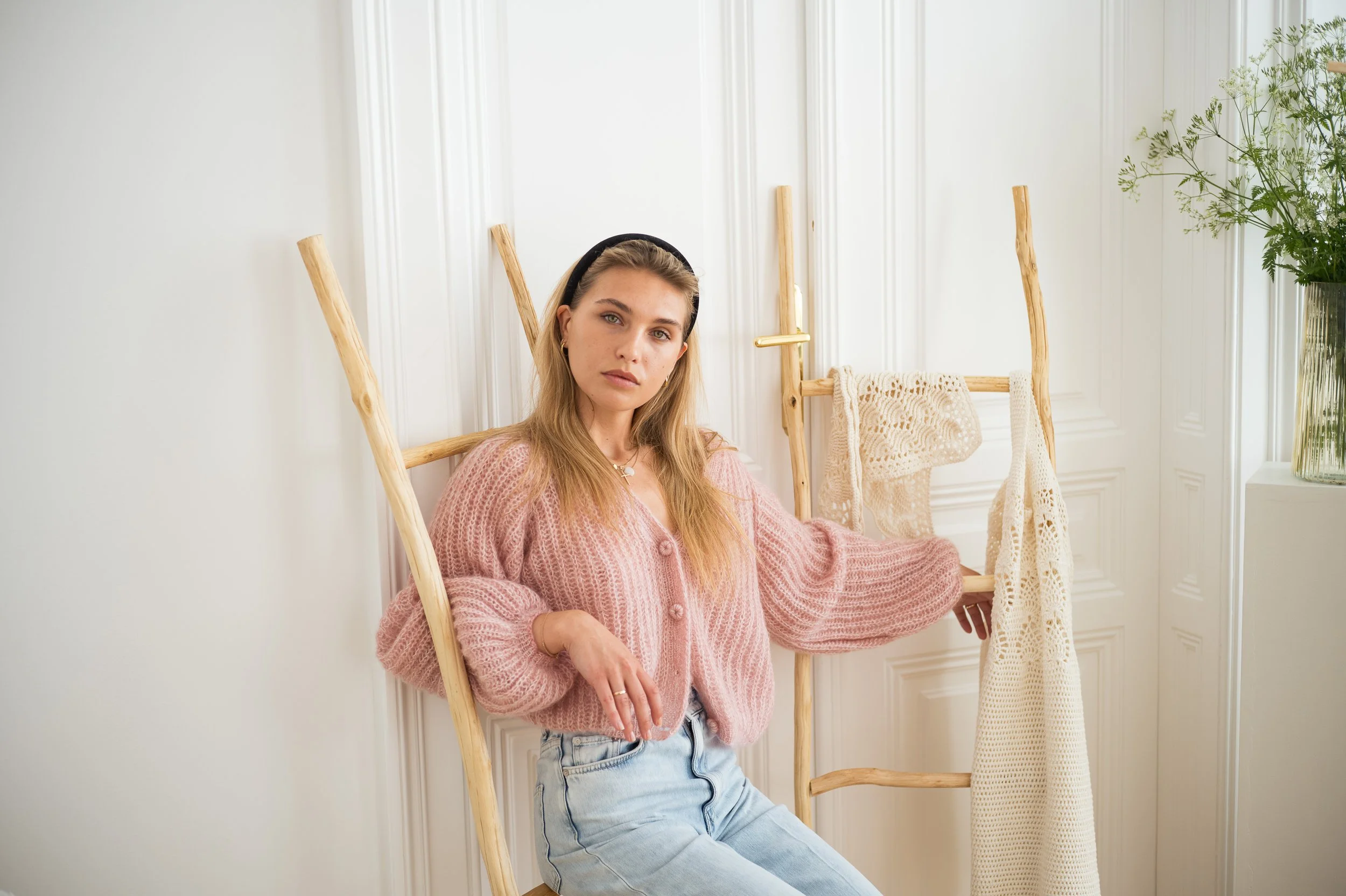 A young woman with blonde hair and blue eyes, wearing a pink sweater with a necklace, poses indoors against a white paneled wall with a wooden ladder, knitted throw, and a potted plant in the background.