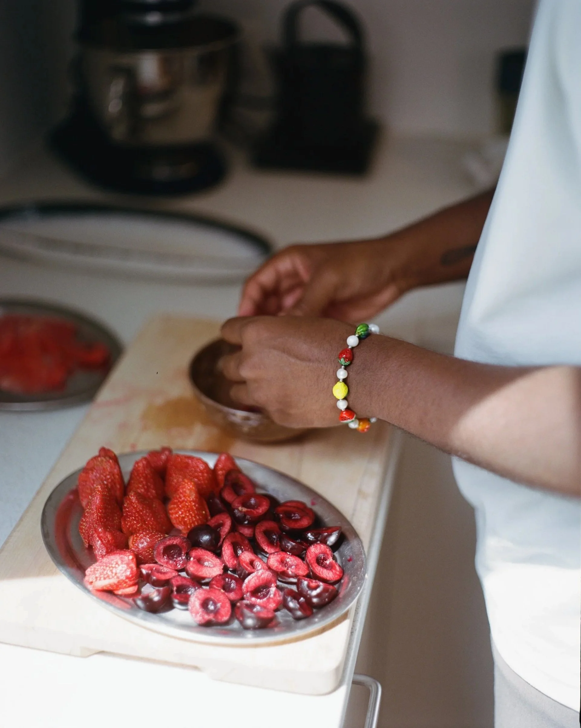 Close-up of a person preparing fresh strawberries and cherries on a kitchen counter, with a bowl of strawberries and cherries in front of them and kitchen appliances in the background.