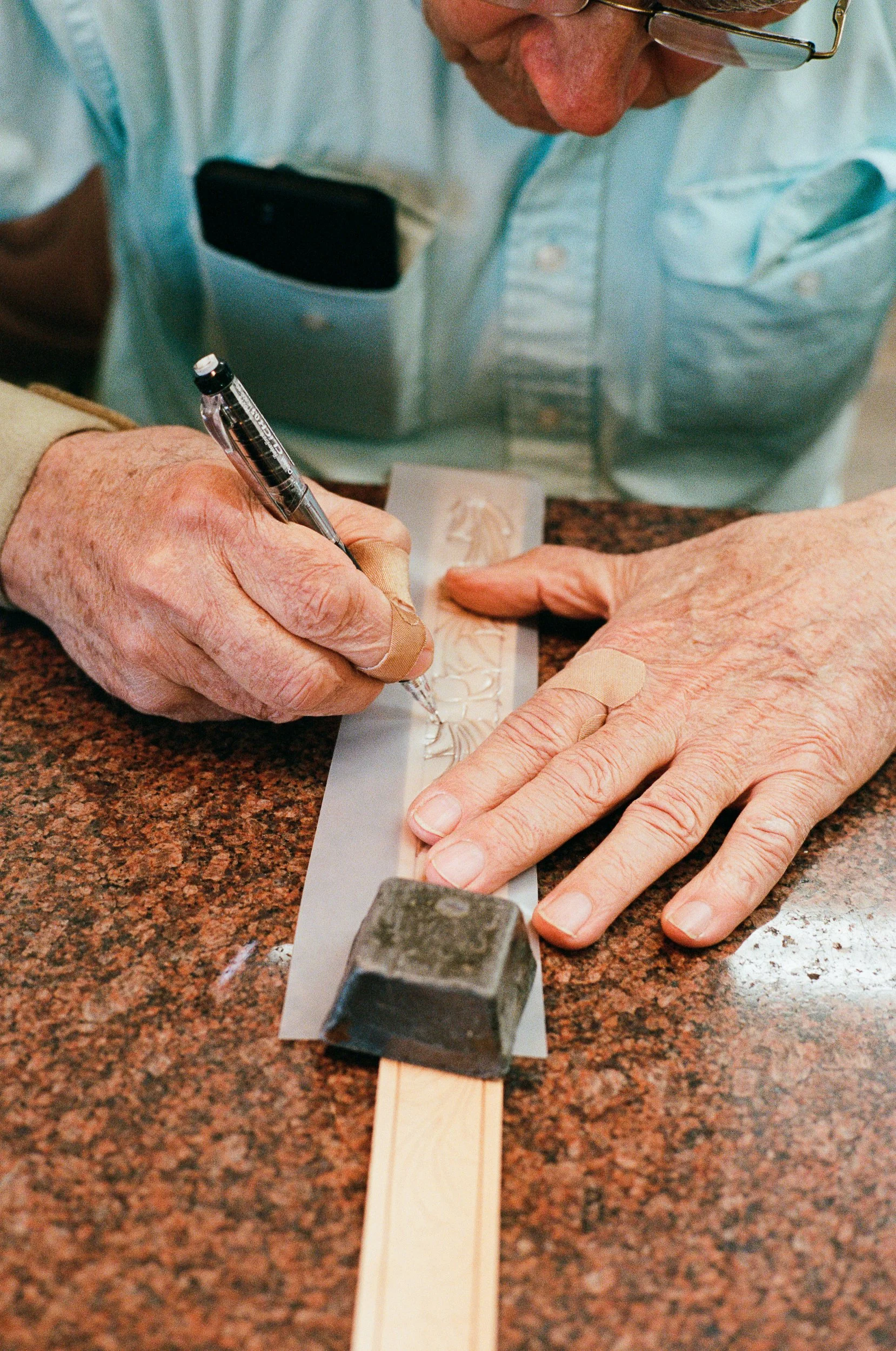 Close-up of an older person's hands engraving a design on a leather belt with a hand tool, while another hand steadies the material on a granite countertop.