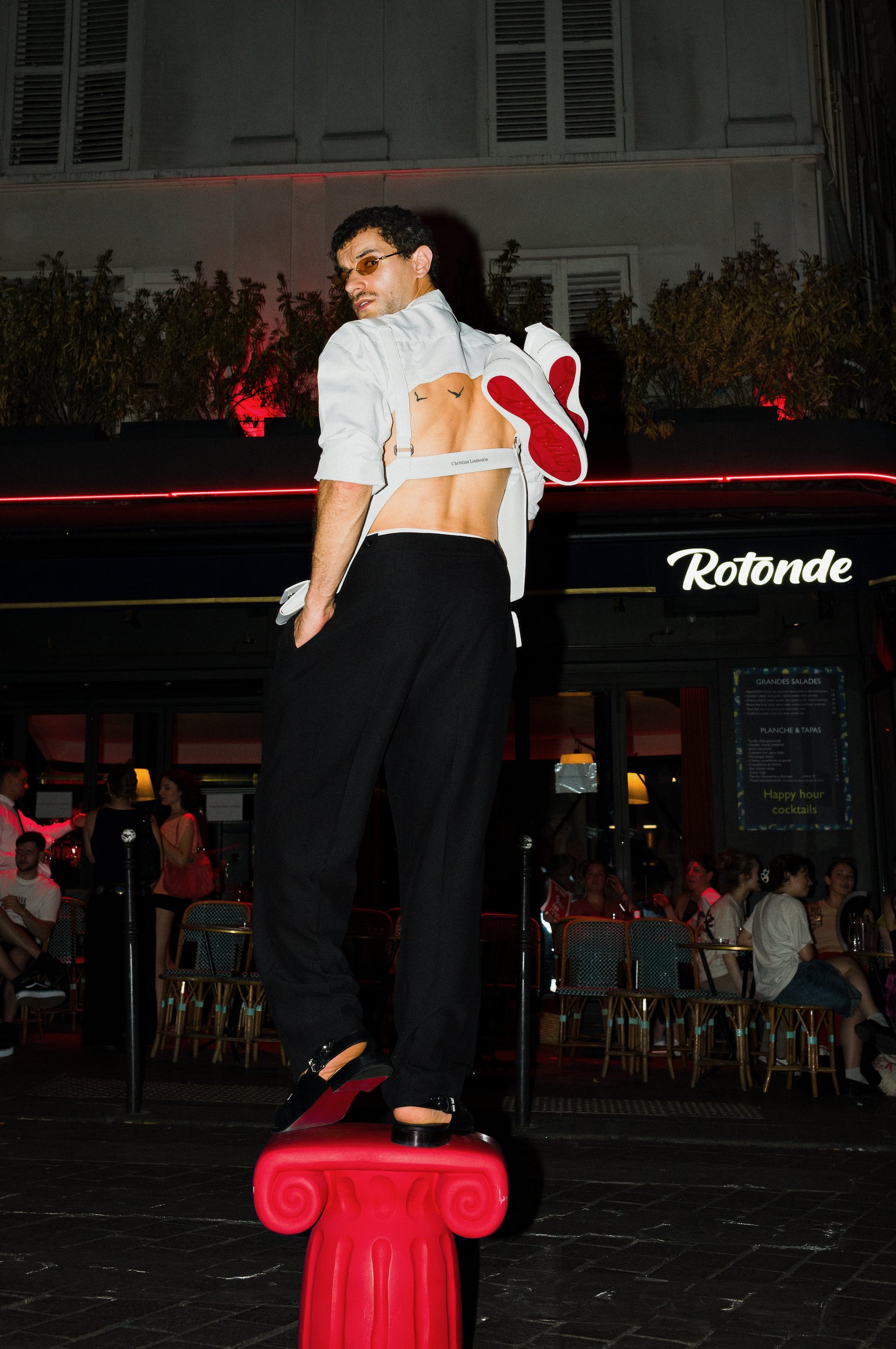 A man with glasses, shirt open revealing his back with two black birds tattooed on it, balancing on a red decorative stool in a lively indoor setting with many people.