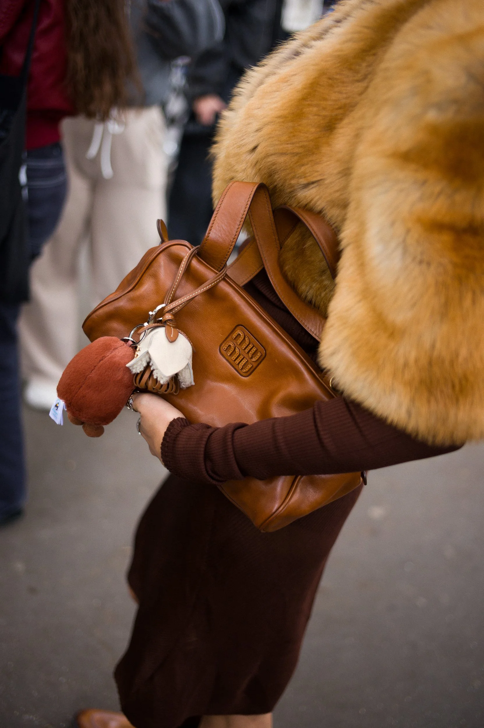 At Paris Fashion Week, A person holding a brown leather handbag with stuffed animal keychains attached, wearing a brown long sleeve shirt and a fur collar or scarf, standing among a crowd.