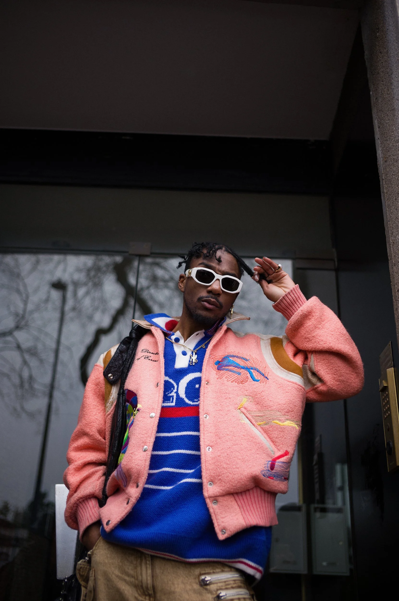 A young person with dark skin, short curly hair, and white sunglasses, standing outdoors near a building, wearing a colorful jacket with various patterns and a blue striped shirt, with trees and windows in the background.