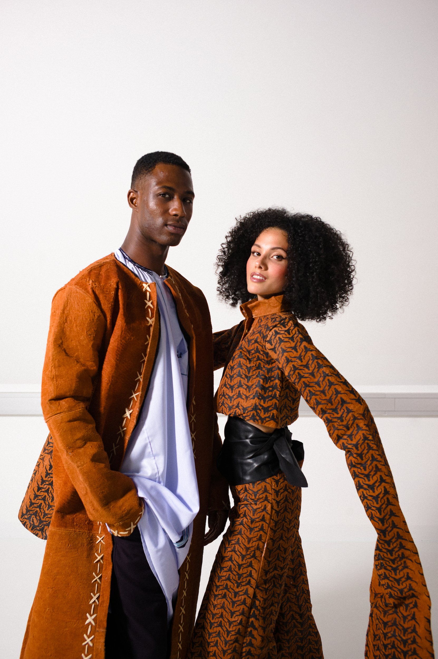 A man and woman standing against a plain white background, both wearing stylish outfits with animal and geometric patterns in warm tones of orange and black.
