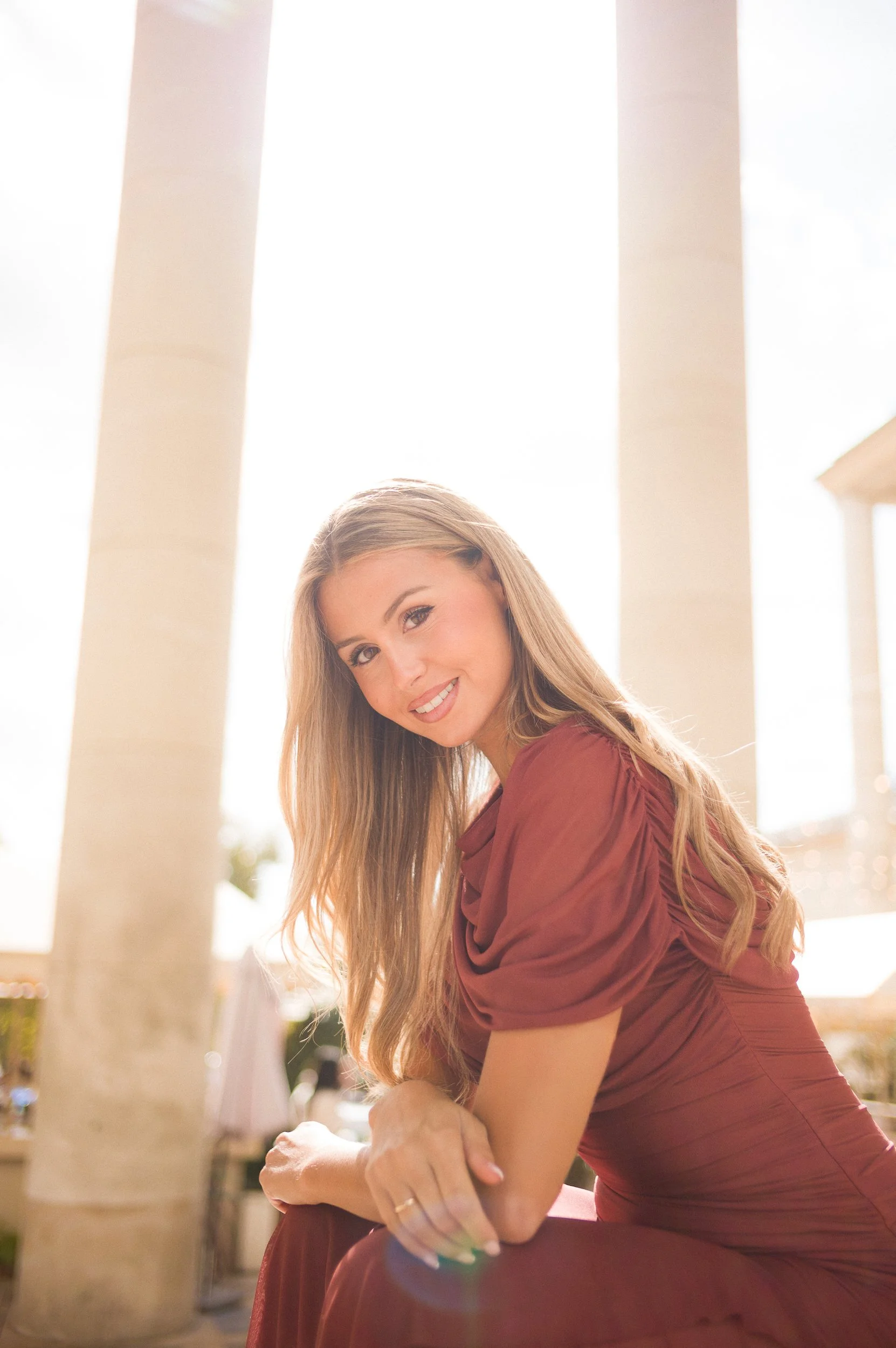 At Paris Fashion Week, A woman with long blonde hair wearing a reddish-brown dress, smiling and looking at the camera, sitting outdoors near large columns with sunlight in the background.