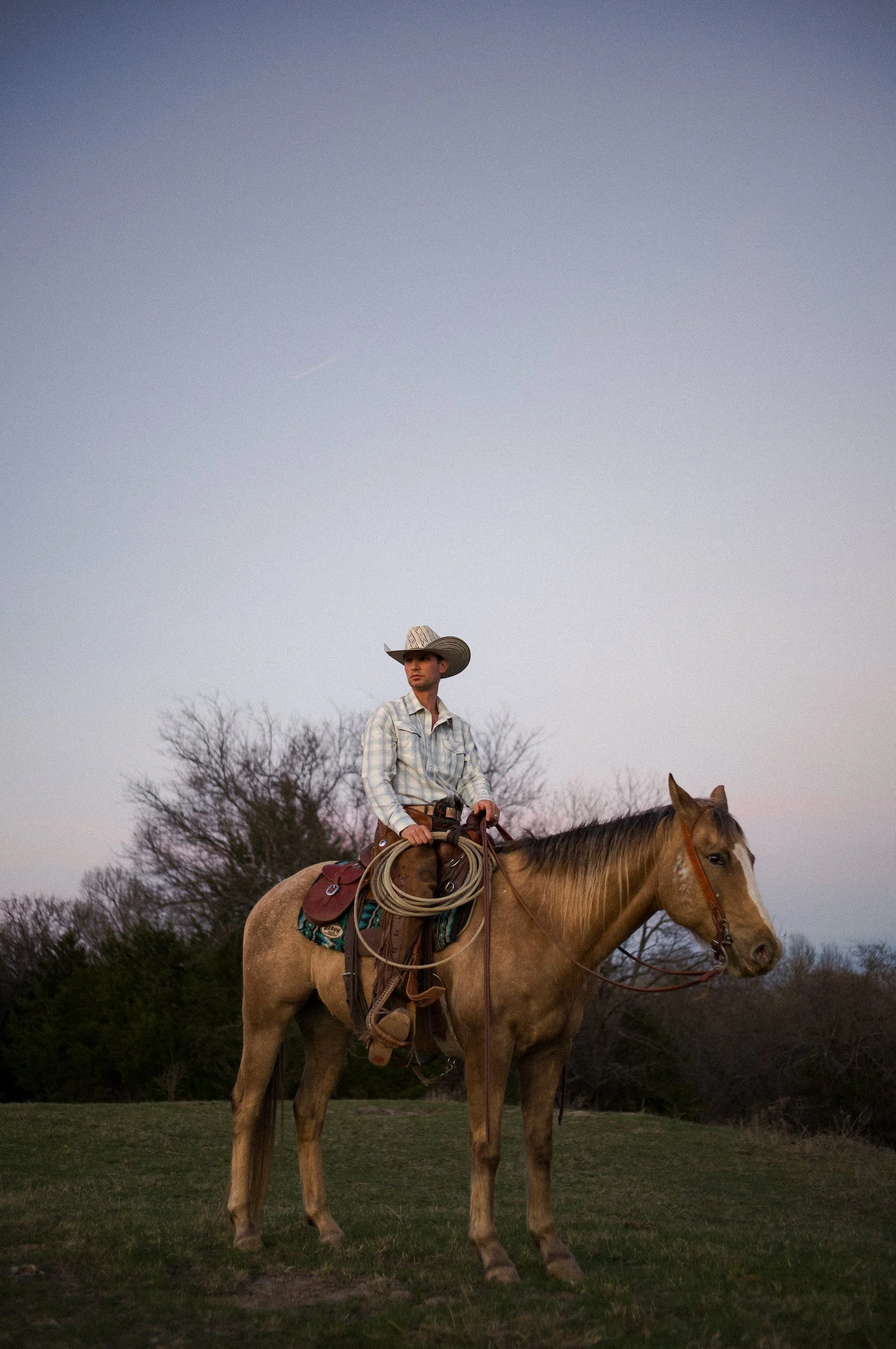 A young man wearing a cowboy hat and plaid shirt riding a tan horse with a saddle and rope, standing on grass with leafless trees and a cloudy sky in the background.