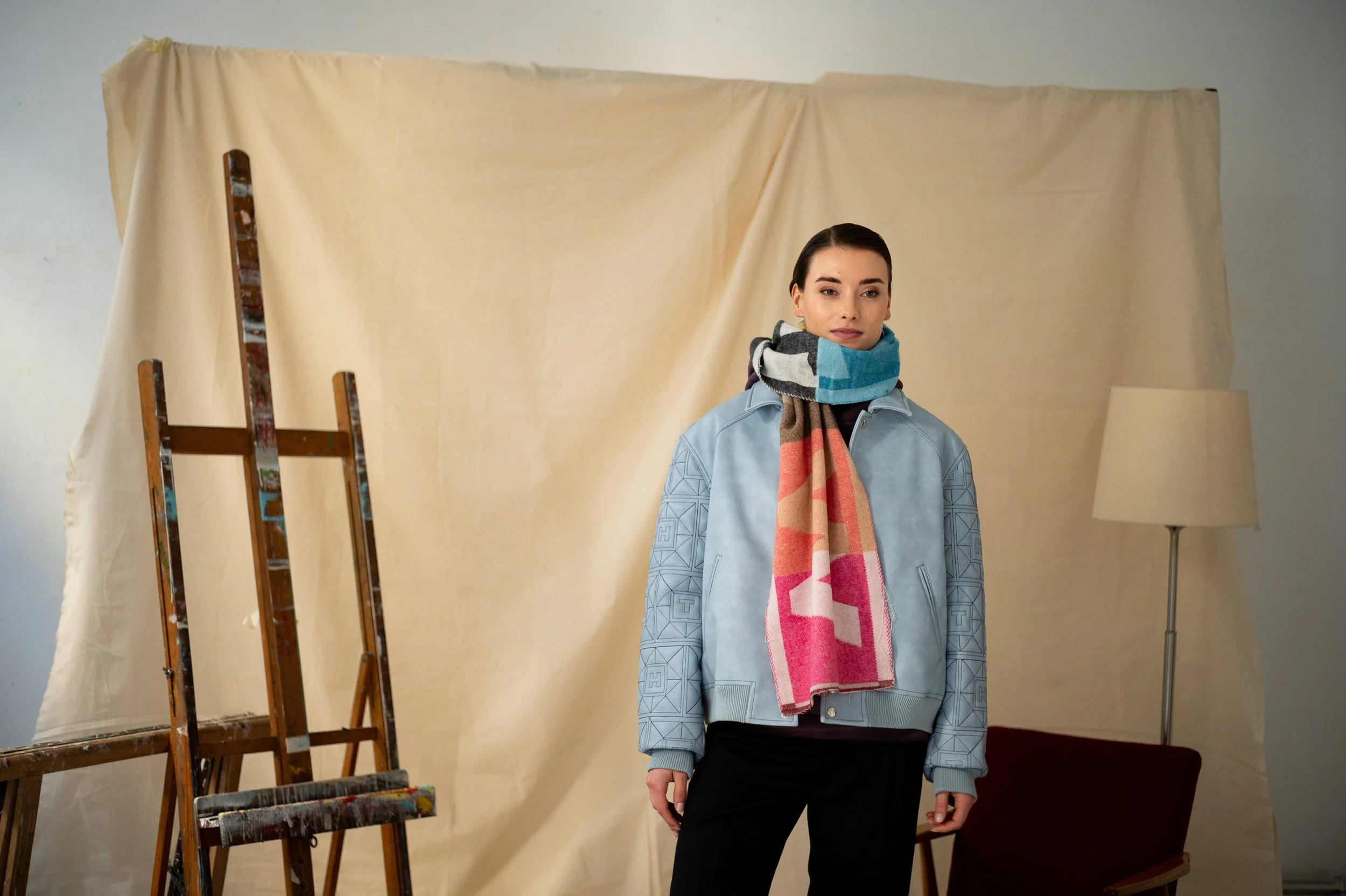 A woman standing indoors in front of a beige backdrop, wearing a blue jacket, a colorful scarf, and black pants, with a painting easel and a lamp nearby.