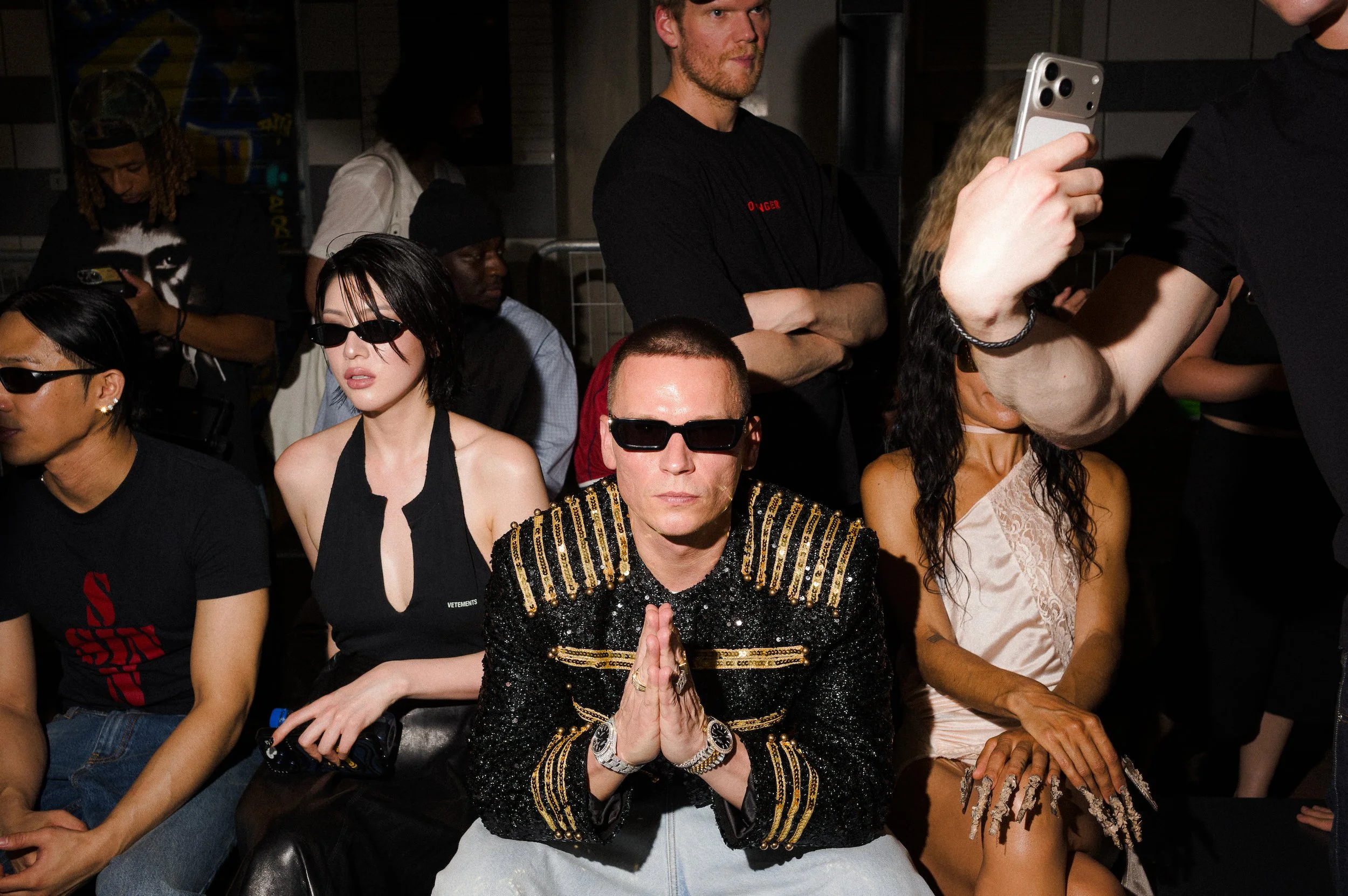 A person in the front is seated with hands pressed in a prayer position, wearing black and gold clothing and sunglasses, with others seated around them, some wearing sunglasses and casual attire, and some standing in the background.