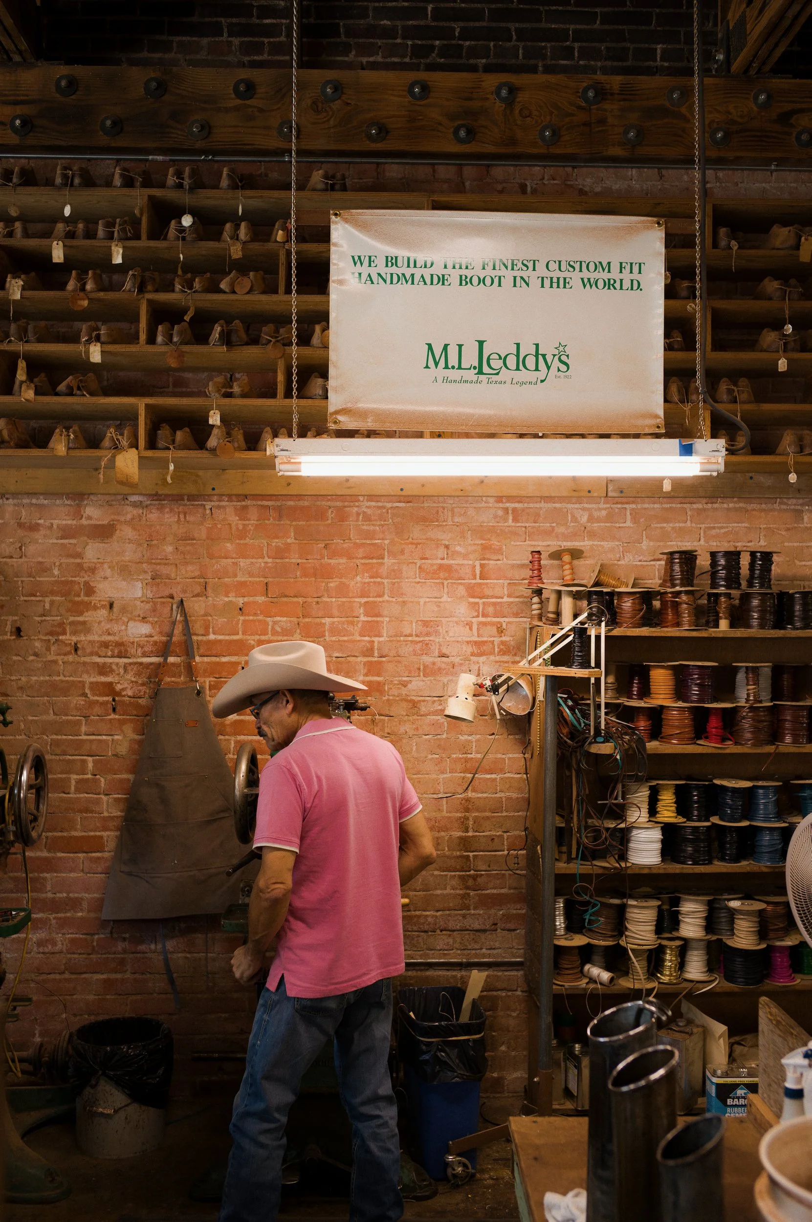 A man in a cowboy hat and pink shirt working in a workshop with shelves of spools of thread and leather materials, and a sign that reads "We build the finest custom fit handmade boot in the world" at M.L. Leddys.