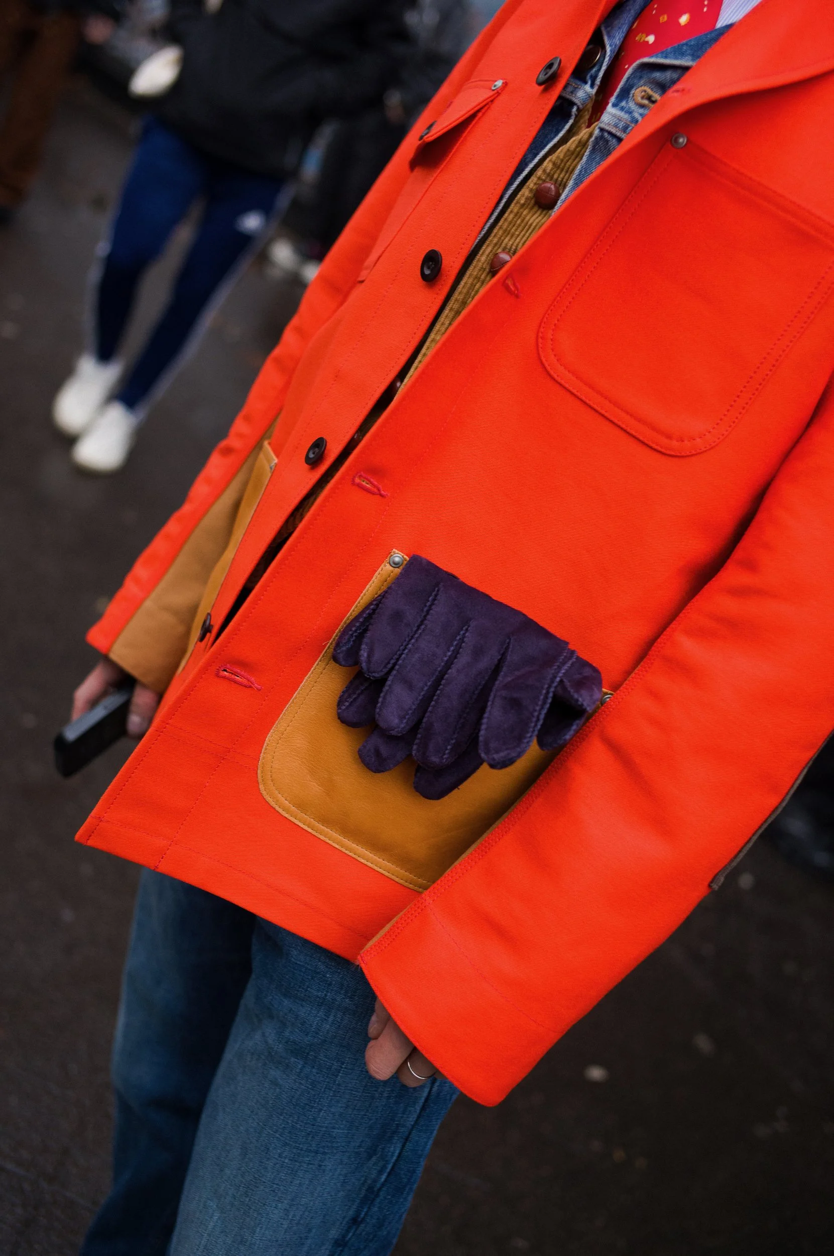 At Paris Fashion Week, Close-up of a person wearing an orange jacket, blue jeans, and a clipboard with a yellow pouch and a pair of purple gloves attached, standing outdoors.
