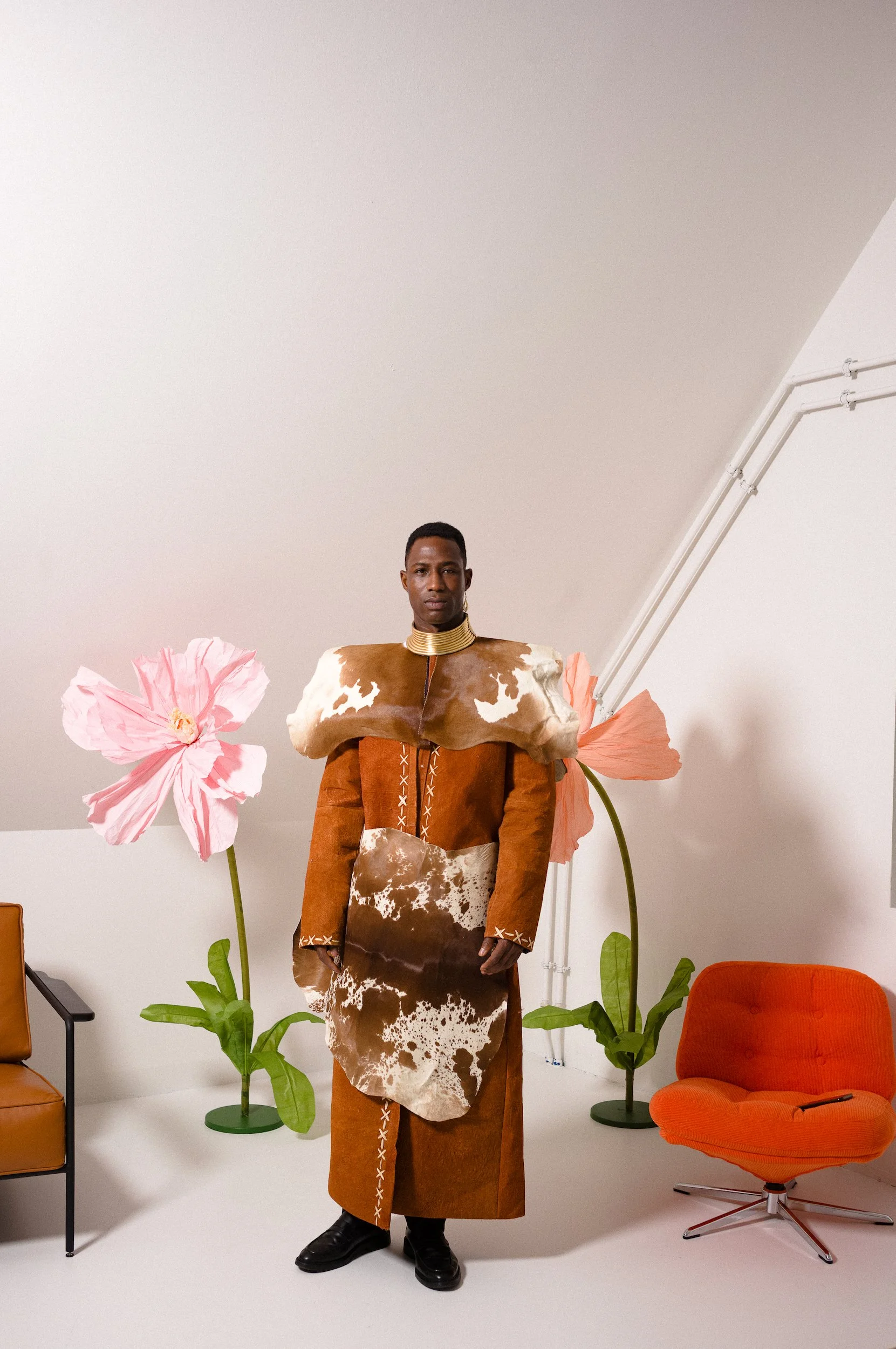 Person standing in a room with large pink flower props, green potted plants, and orange chairs. They are wearing a brown and white patterned outfit with a cowhide motif, and a gold choker. The background shows a plain wall and partial ceiling.