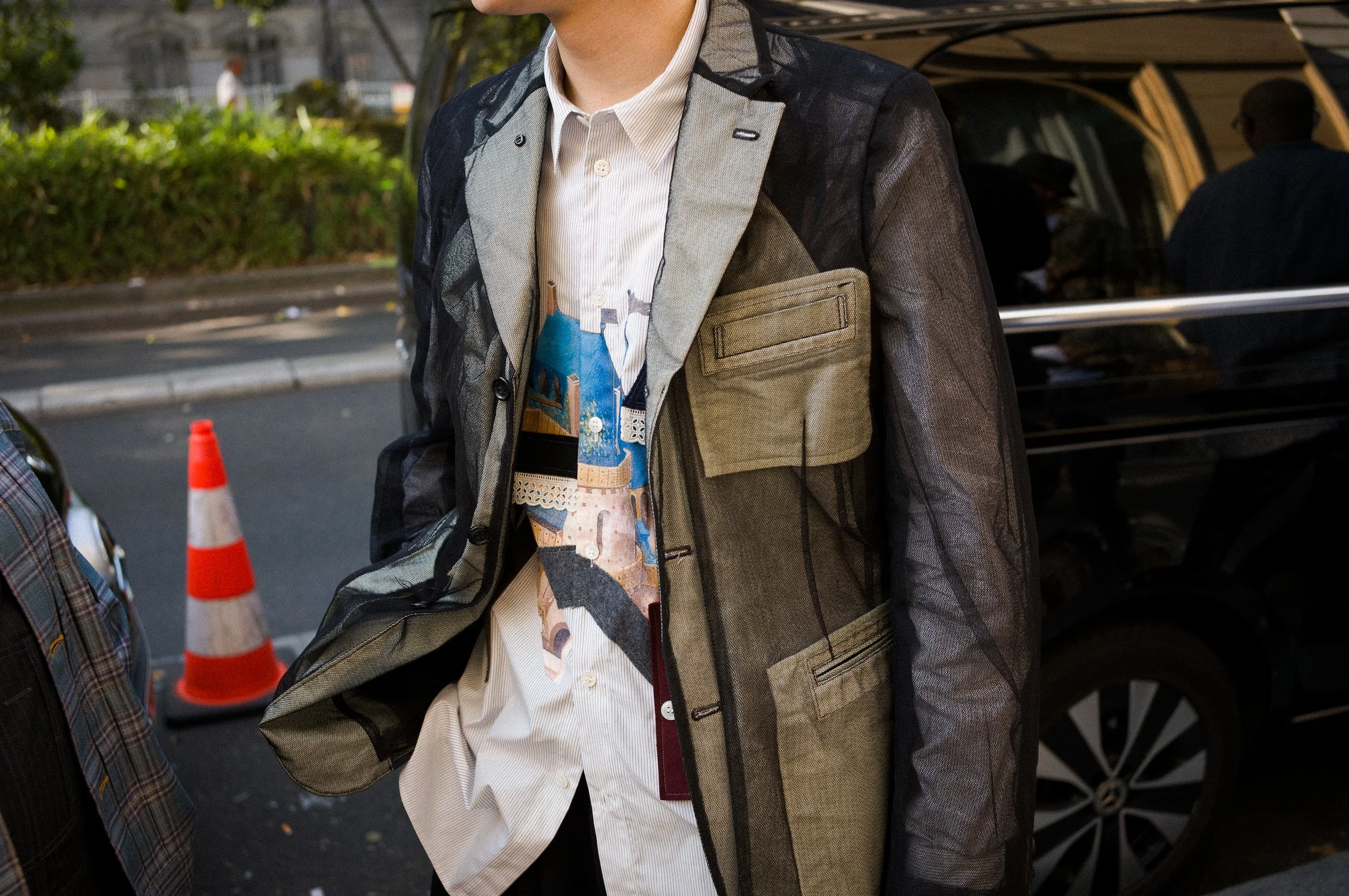 Close-up of a person dressed in layered clothing, including a printed shirt, a light jacket with a pocket, and a transparent mesh outer layer, standing outdoors near a vehicle with other people and a traffic cone in the background.