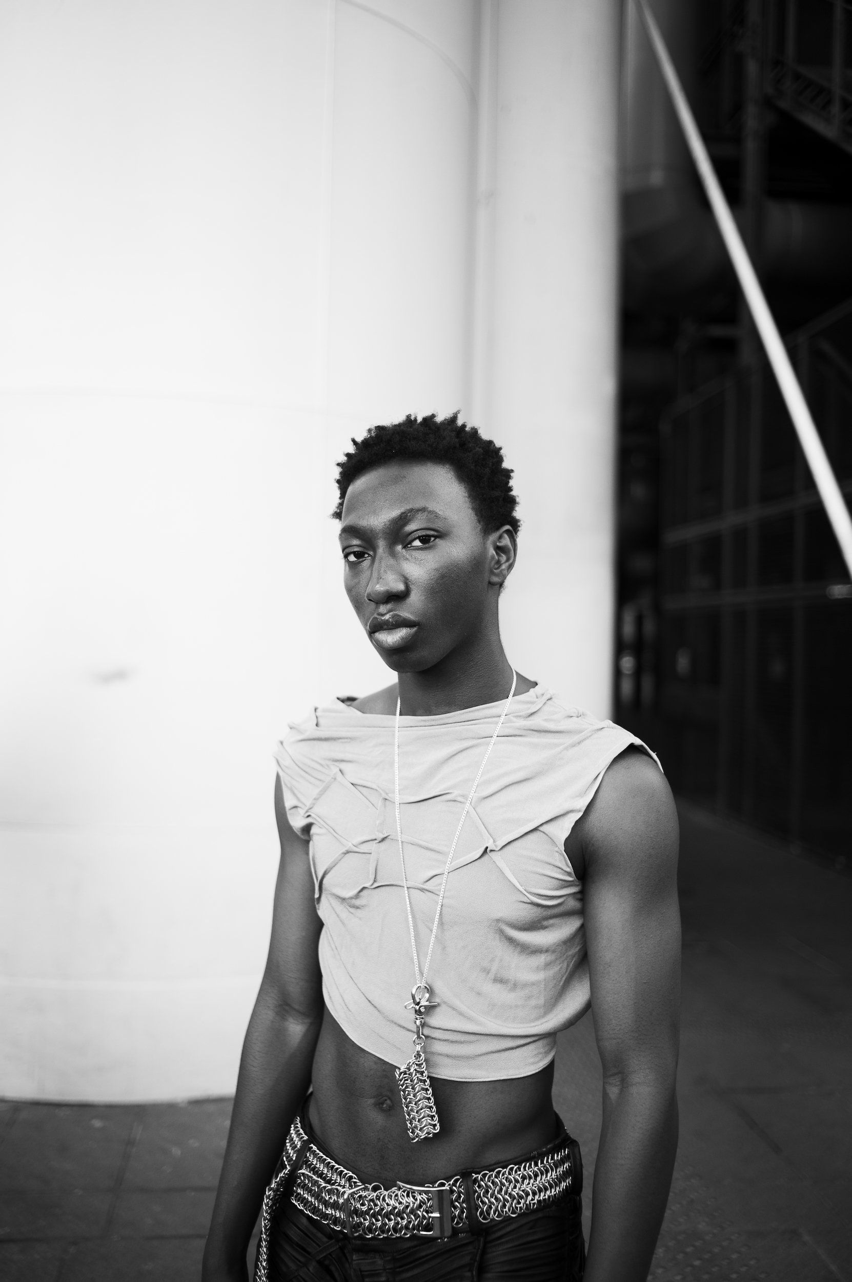 At Paris Fashion Week, Black and white photo of a man with short hair wearing a ripped top, a chain necklace, and a chain belt, standing inside a building with industrial background.