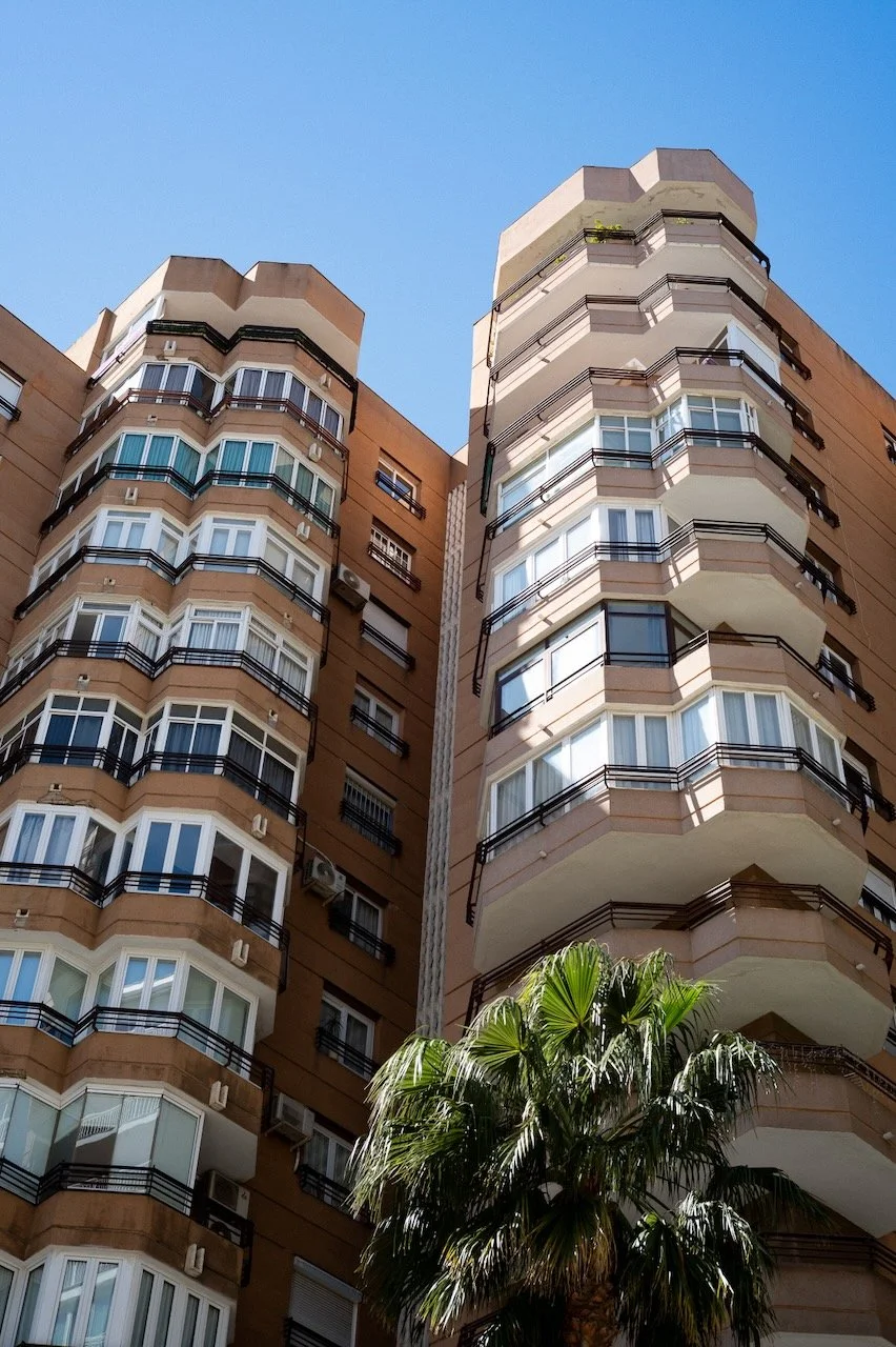 In Spain, A tall apartment building with multiple floors, balconies, and windows, with a palm tree in the foreground against a clear blue sky.