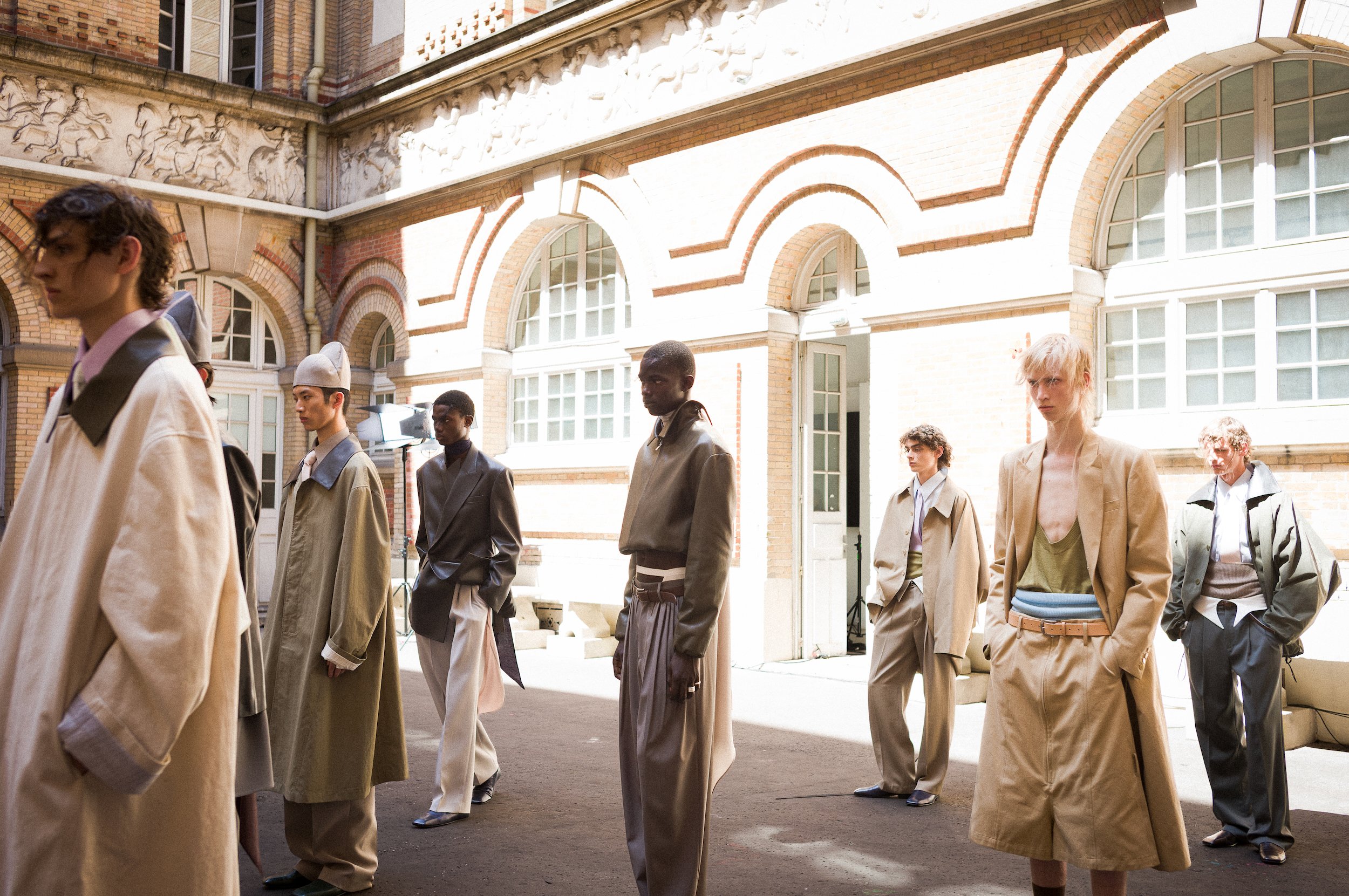 Finale of the Sean Suan show at Paris fashion week. All dressed in fashionable, neutral-colored clothing and sunglasses, walking outdoors near a historic building with arched windows and decorative brickwork.