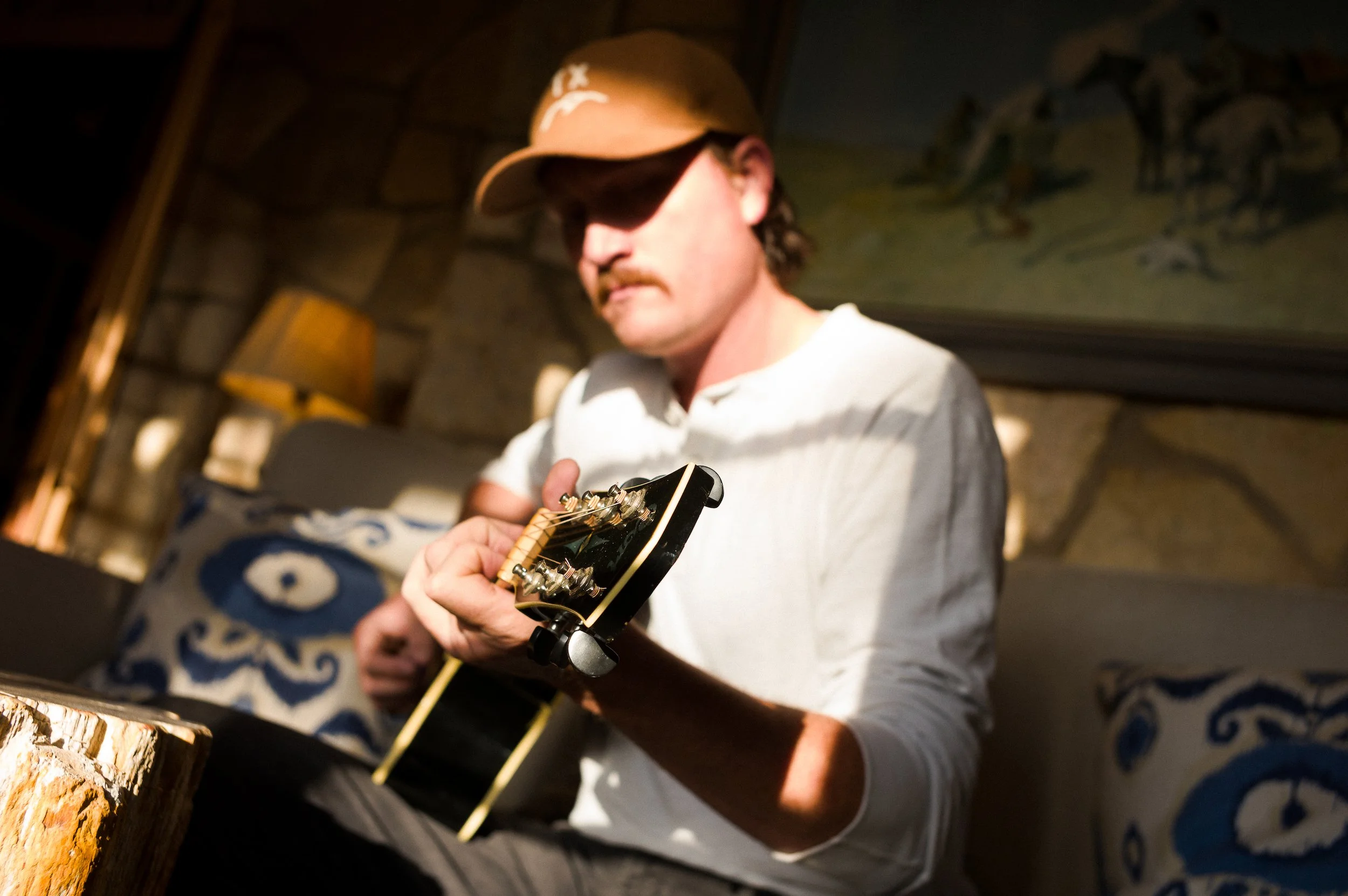 A man wearing a brown cap with sunglasses and a white shirt is playing an electric guitar in a cozy indoor setting with wooden walls and a decorative blue and white patterned pillow.
