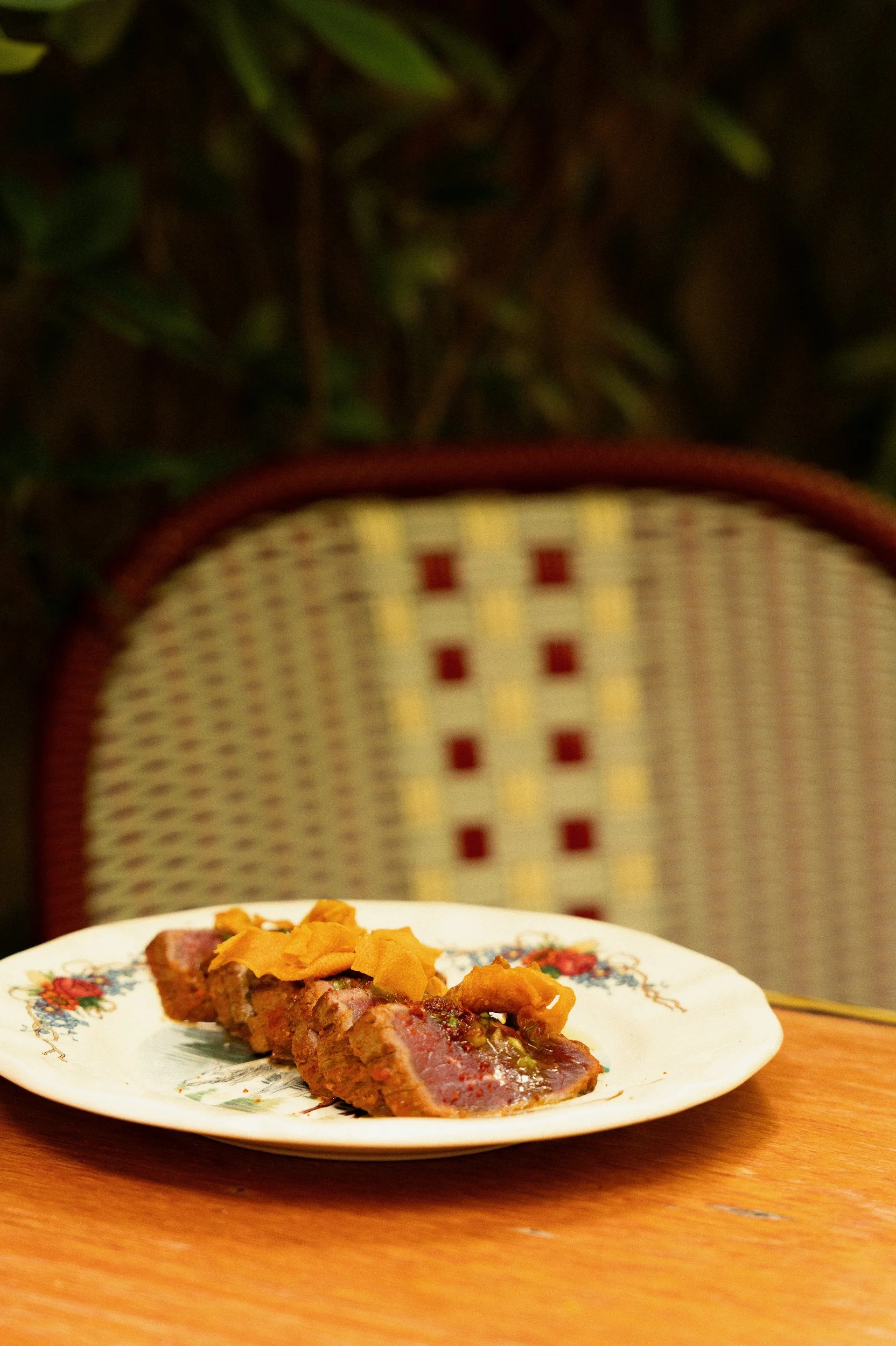 A plate of cooked meat topped with roasted vegetables, placed on a wooden table. In the background, a chair with a patterned cushioned backrest and some greenery are visible.