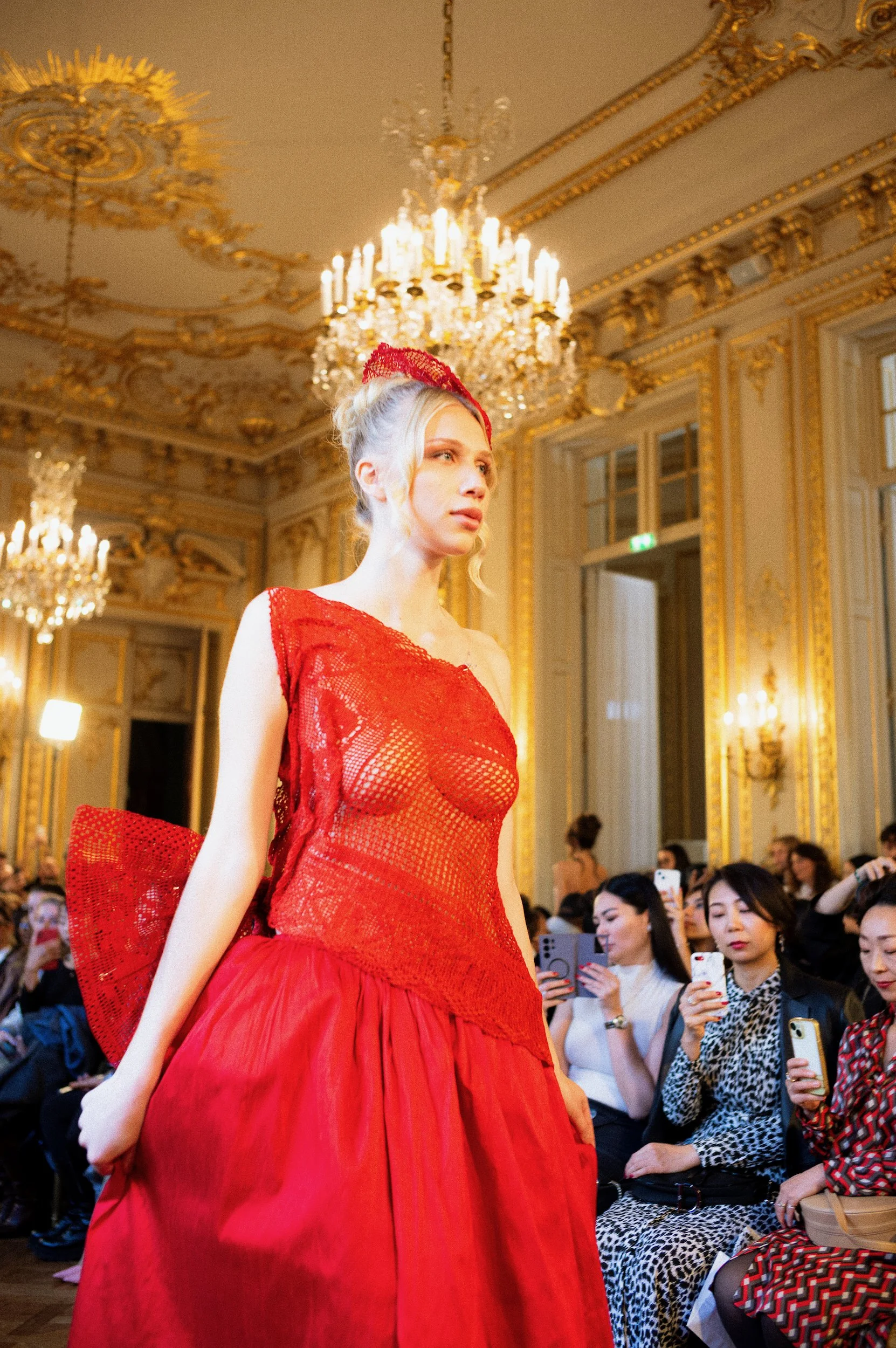 At Paris Fashion Week, A woman is walking on a runway in an ornate room with golden decorations and chandeliers, wearing a red fashion design.