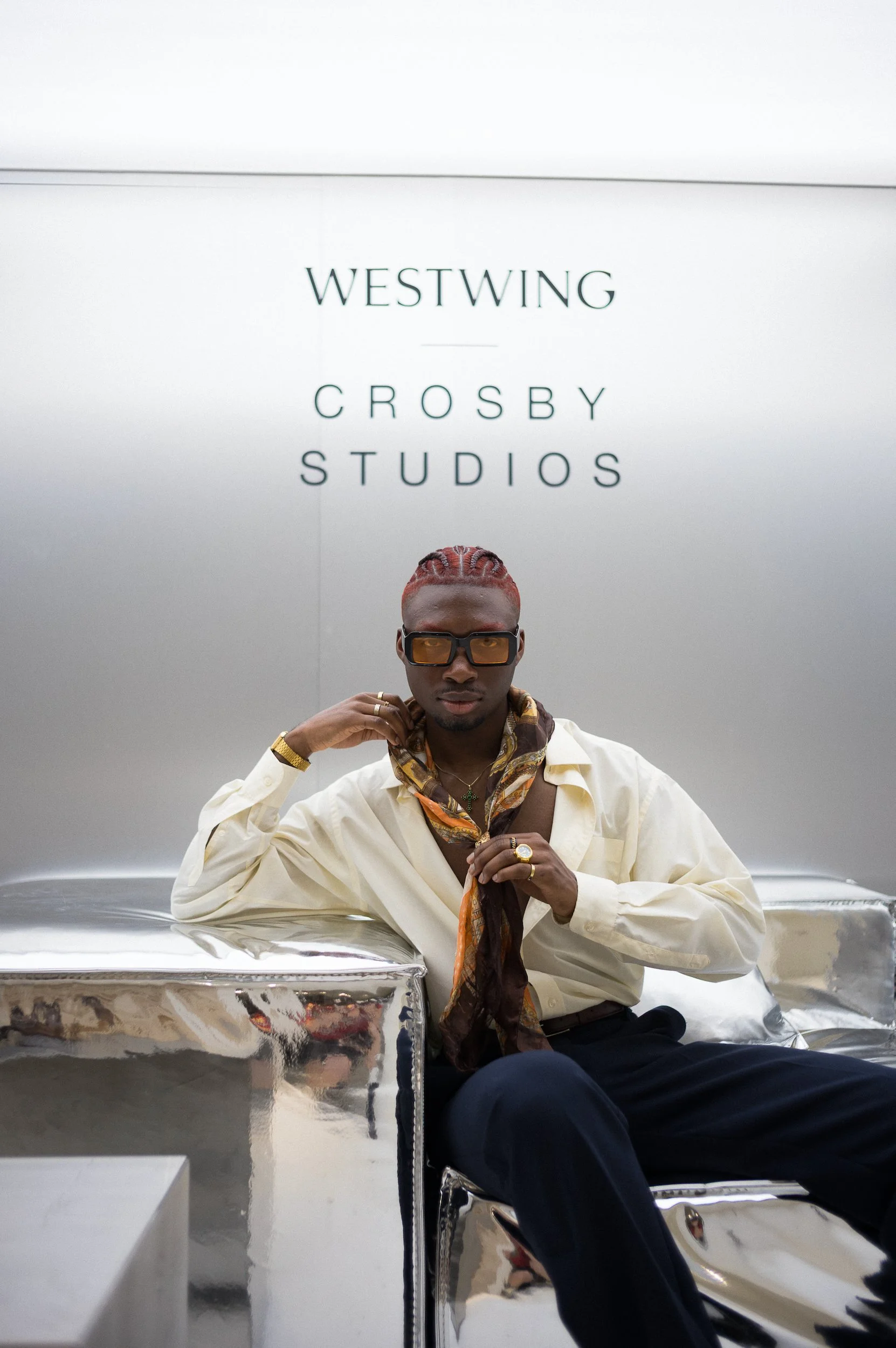 At Paris Fashion Week, A man with red hair, wearing sunglasses and multiple jewelry pieces, relaxing on a shiny metallic bench in front of a white wall with the text 'Westwing Crosby Studios'.