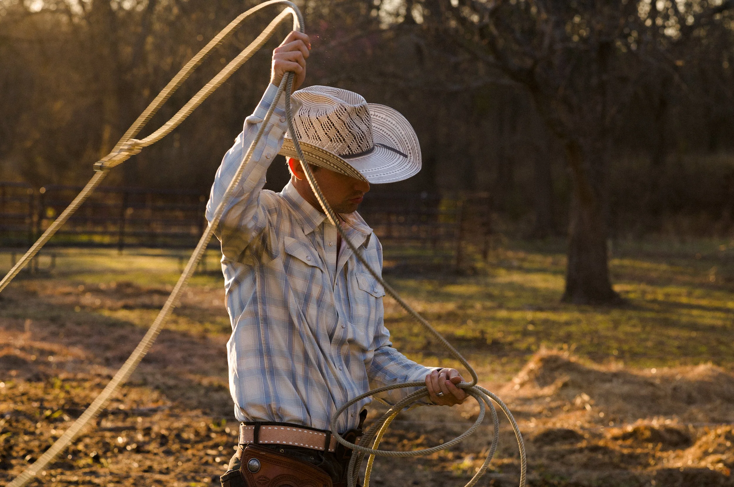 A man wearing a plaid shirt, cowboy hat, and cowboy boots appears to be roping cattle during sunset or sunrise.