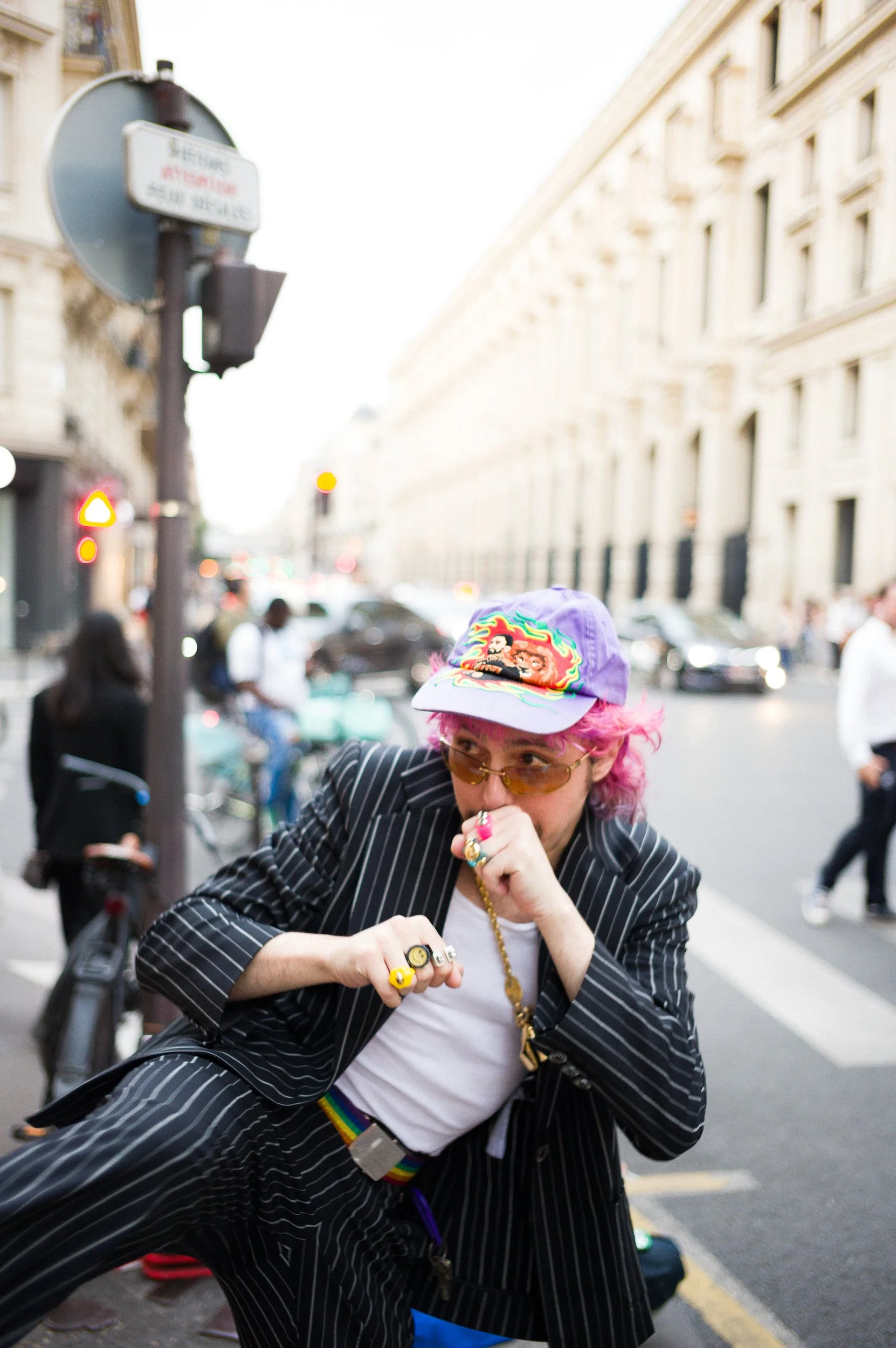 At Paris Fashion Week, A man with pink hair wearing a colorful hat, glasses, gold jewelry, and a black pinstripe suit, posed in a busy city street with buildings, cars, and other pedestrians in the background.