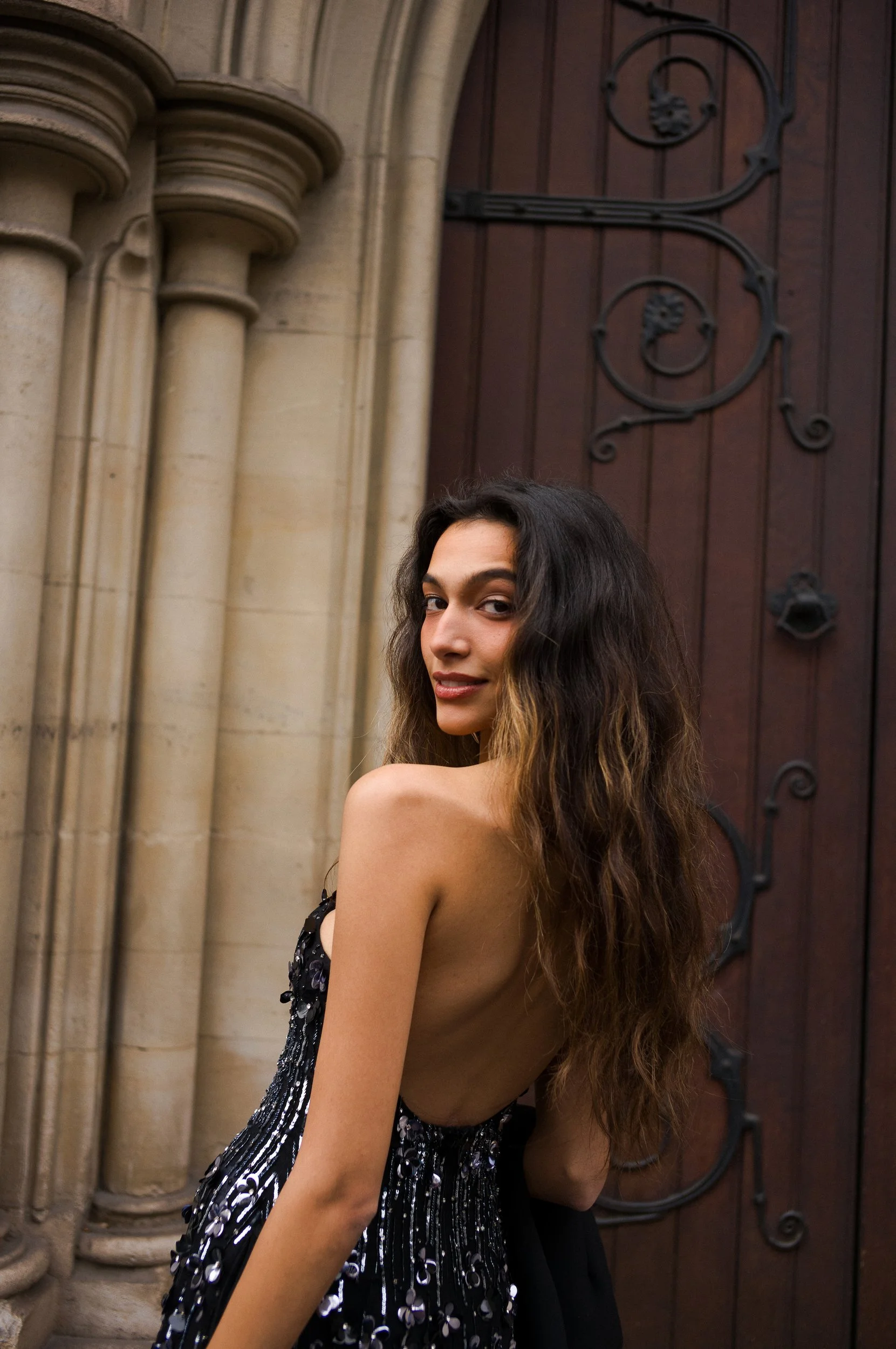 At Paris Fashion Week, A woman with long brunette hair, partially turned, smiling slightly, standing in front of a dark wooden door with decorative black wrought iron hinges, part of a stone building with columns.