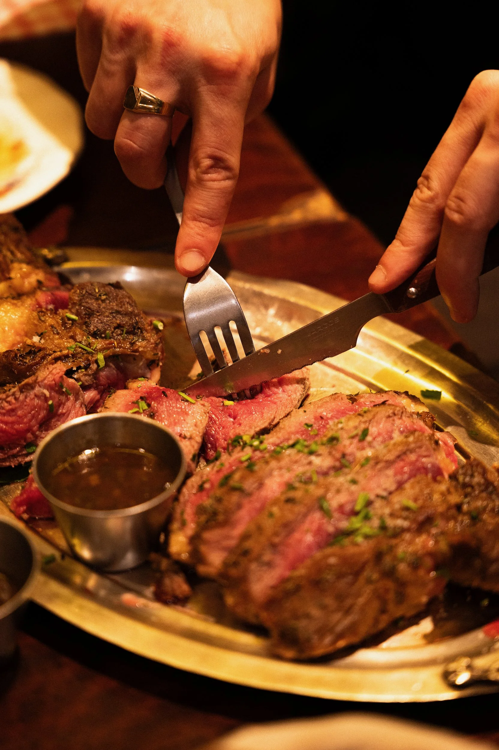 Close-up of a person slicing a medium-rare roast beef or prime rib using a knife and fork, with a small cup of au jus or gravy nearby, on a round wooden serving tray.
