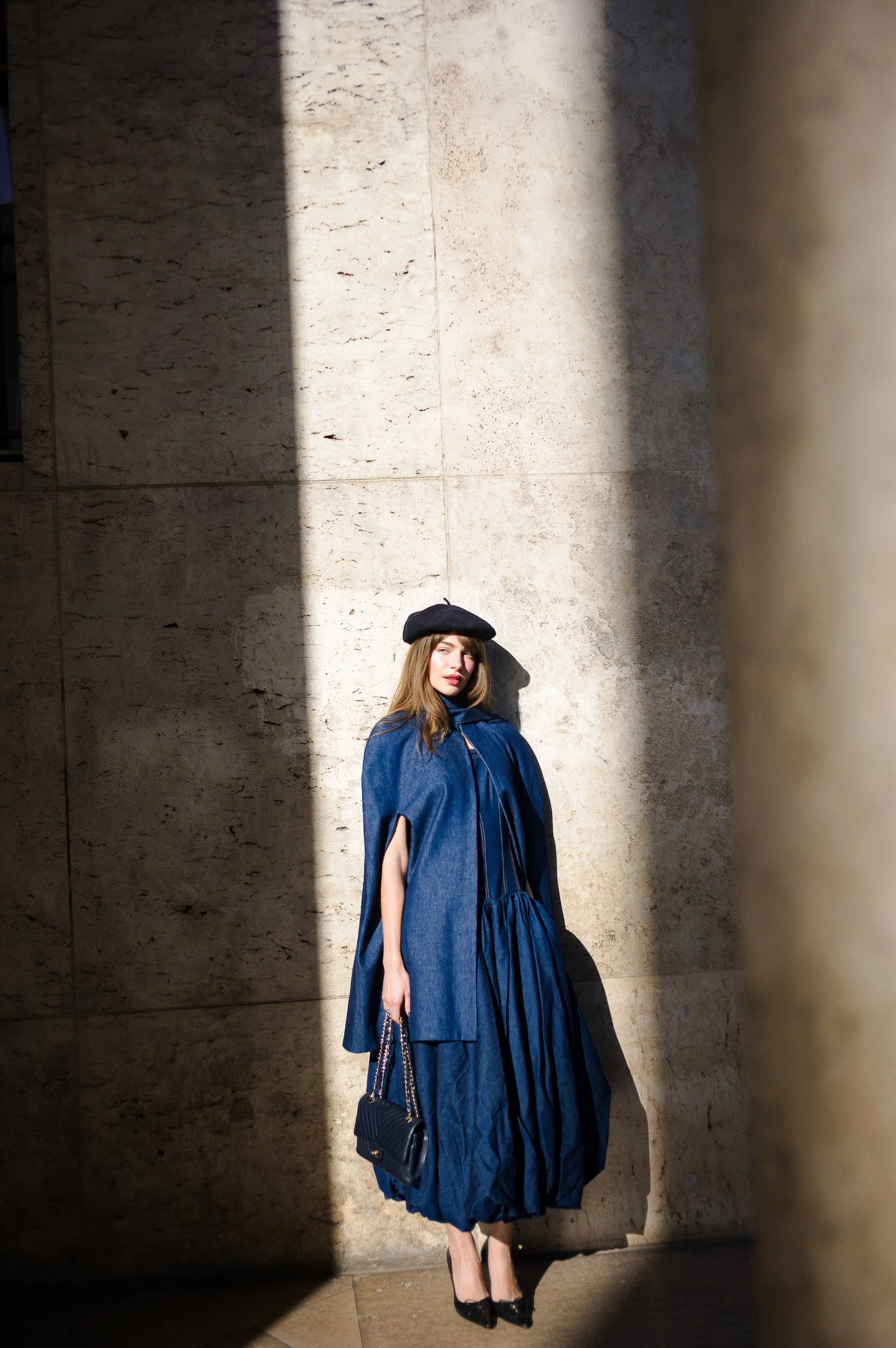 At Paris Fashion Week, A woman in a long blue dress, black heels, a black beret, and carrying a black purse stands against a marble wall. sunlight creates shadowed and highlighted areas on the wall.