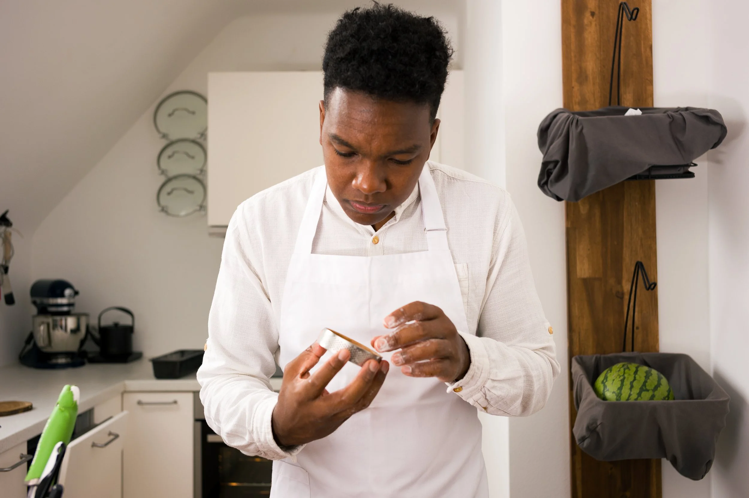 A chef wearing a white shirt and apron is looking down at their hands while holding a kitchen tool in a domestic kitchen.