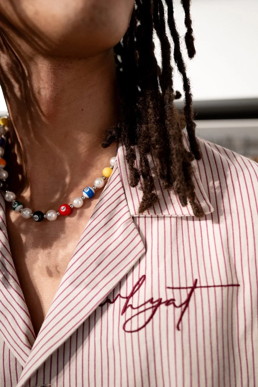 Close-up of a man wearing a striped collared shirt with red and white lines, an embroidered signature-style logo, and a colorful beaded necklace with alphabet and number beads, and dreadlocked hair.