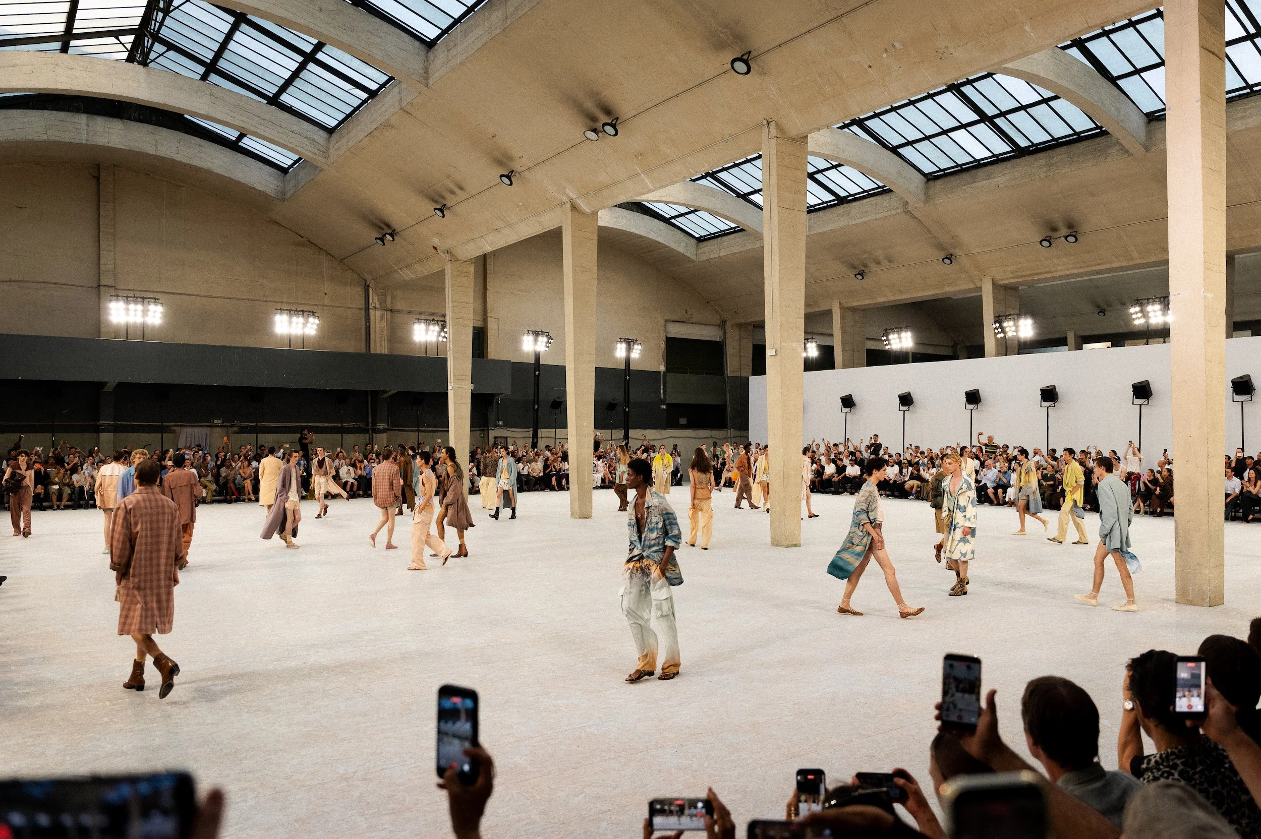 Dries Van Noten Show at Paris Fashion week, the finale walking and sitting inside a large indoor space with high glass ceilings, bright lighting, and concrete supports, with many people recording or taking photos with their phones.