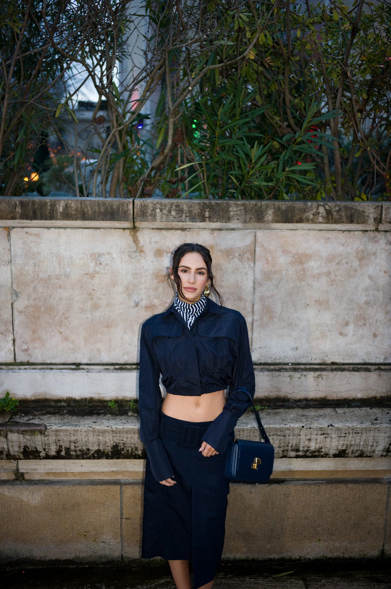 A woman standing outdoors against a concrete wall with green plants in the background. She is wearing a navy jacket, a black skirt with a slit, black gloves, and is carrying a black handbag. Her hair is styled in loose waves and she is looking at the