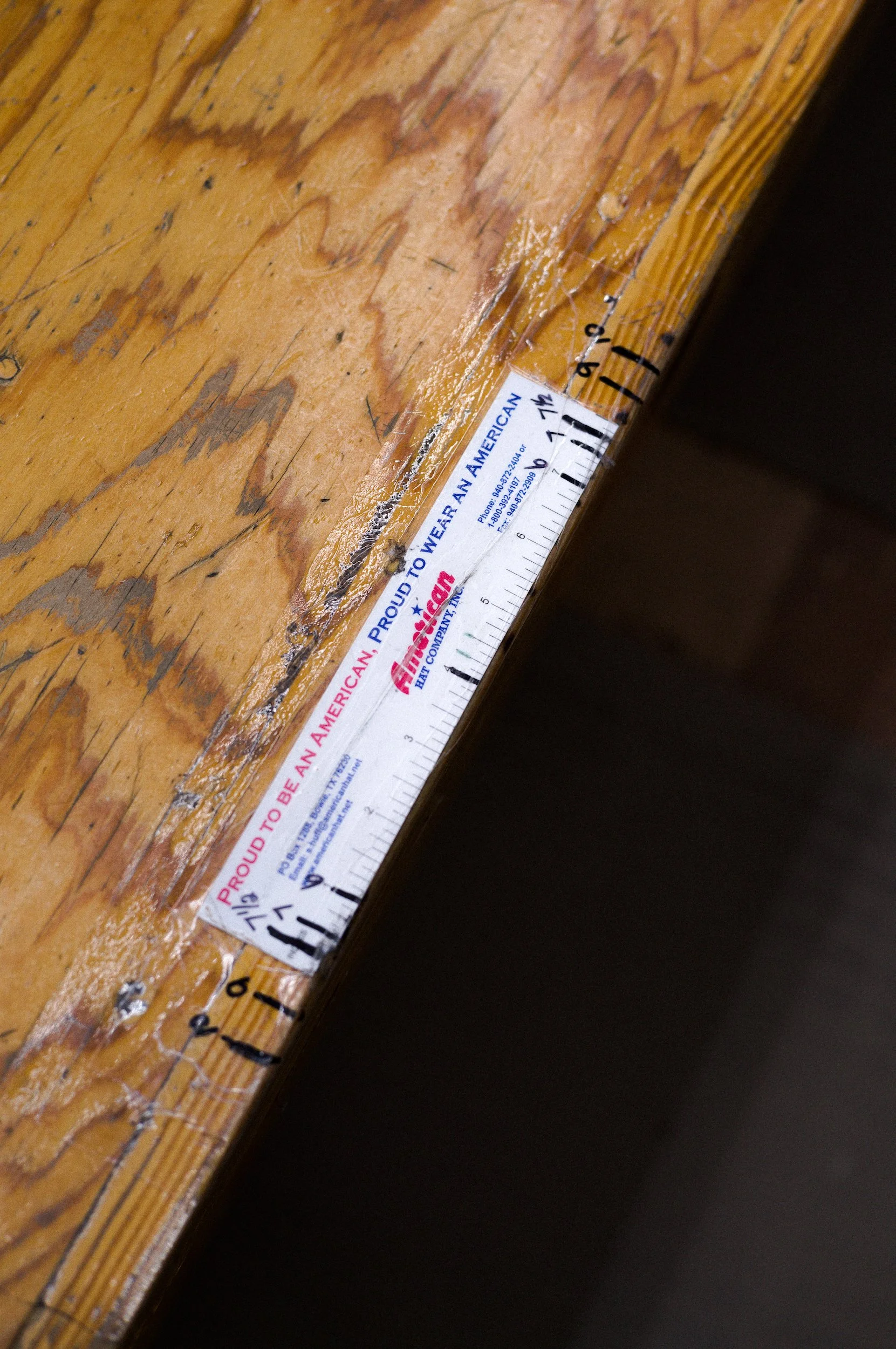 Close-up of a wooden surface with a ruler showing the American hat company logo which is worn and peeling.