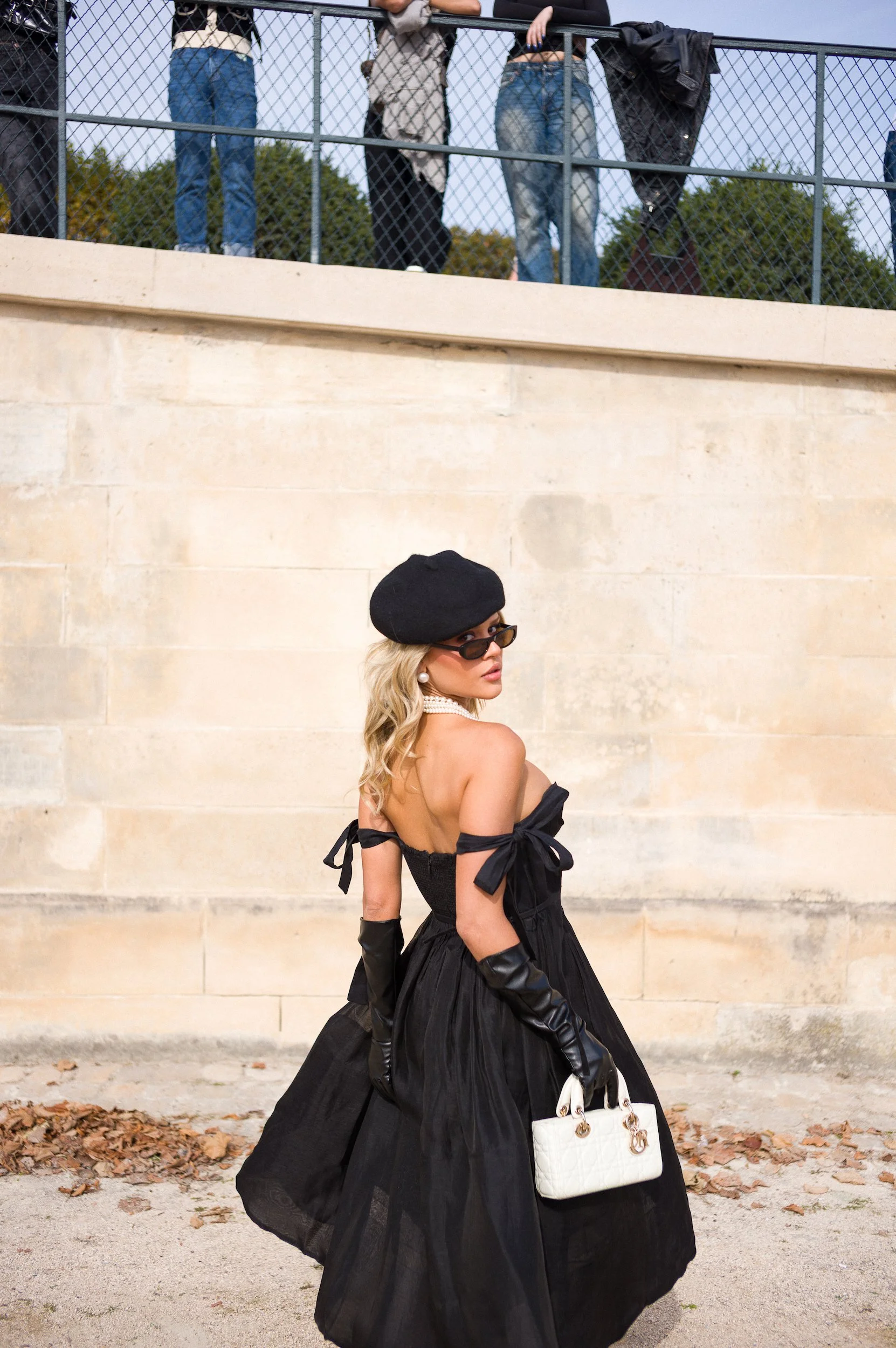 A woman with blonde hair, wearing a black beret, sunglasses, pearl earrings, a pearl necklace, black gloves, a black strapless dress with bow accents on the shoulders, and carrying a white handbag, standing outdoors against a beige stone wall with fa