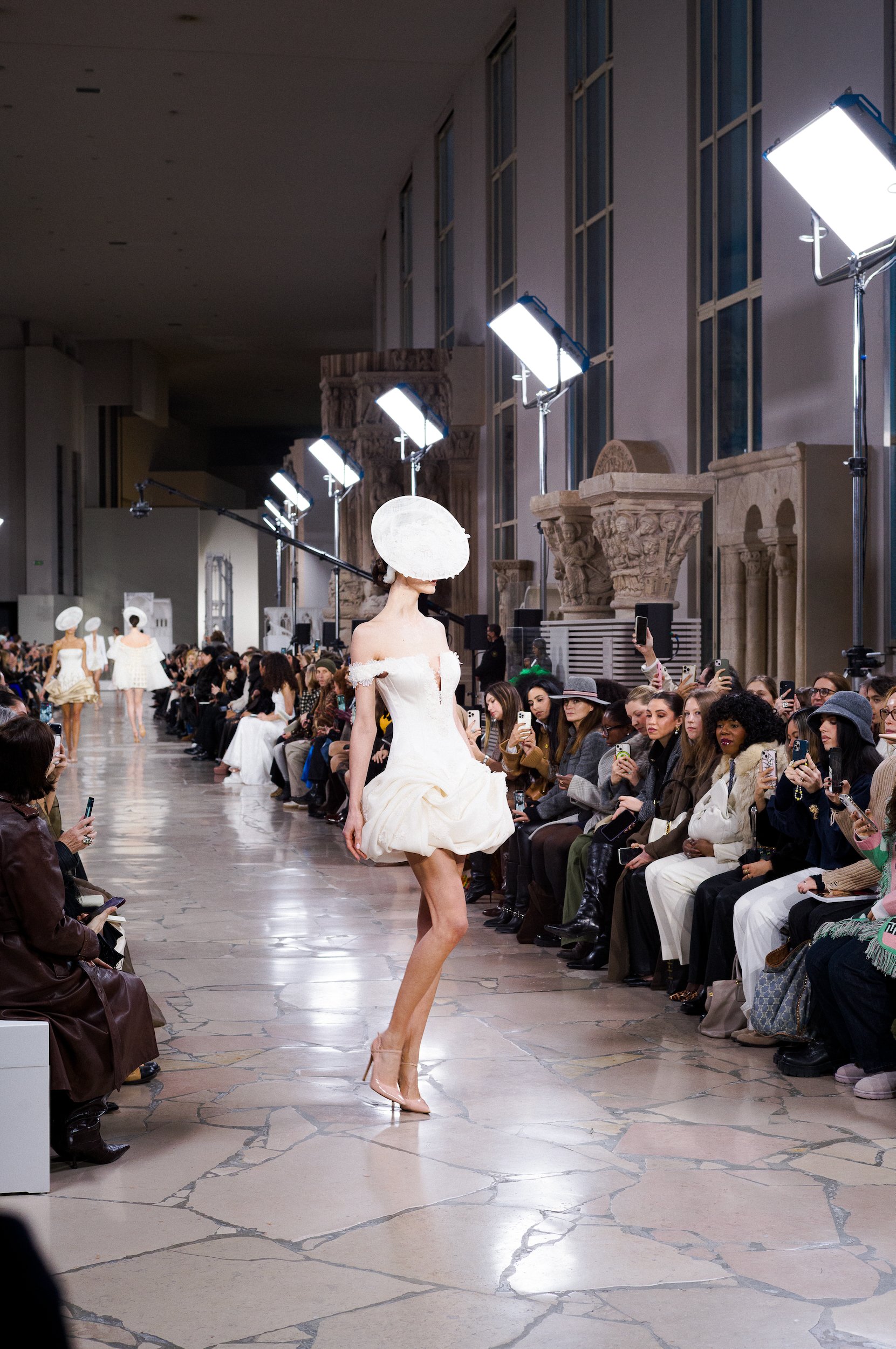 At Paris Fashion Week, Fashion runway show with models walking on runway, audience seated on both sides, large windows and classical columns in background, bright lighting.