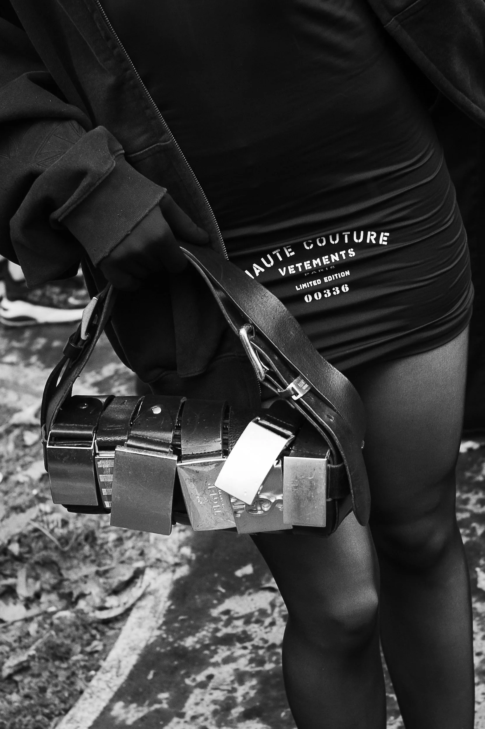At Paris Fashion Week, Close-up of a person's black outfit, including a jacket, top, and leggings, holding multiple large padlocks in their hand, with a background of a parking lot and fallen leaves.