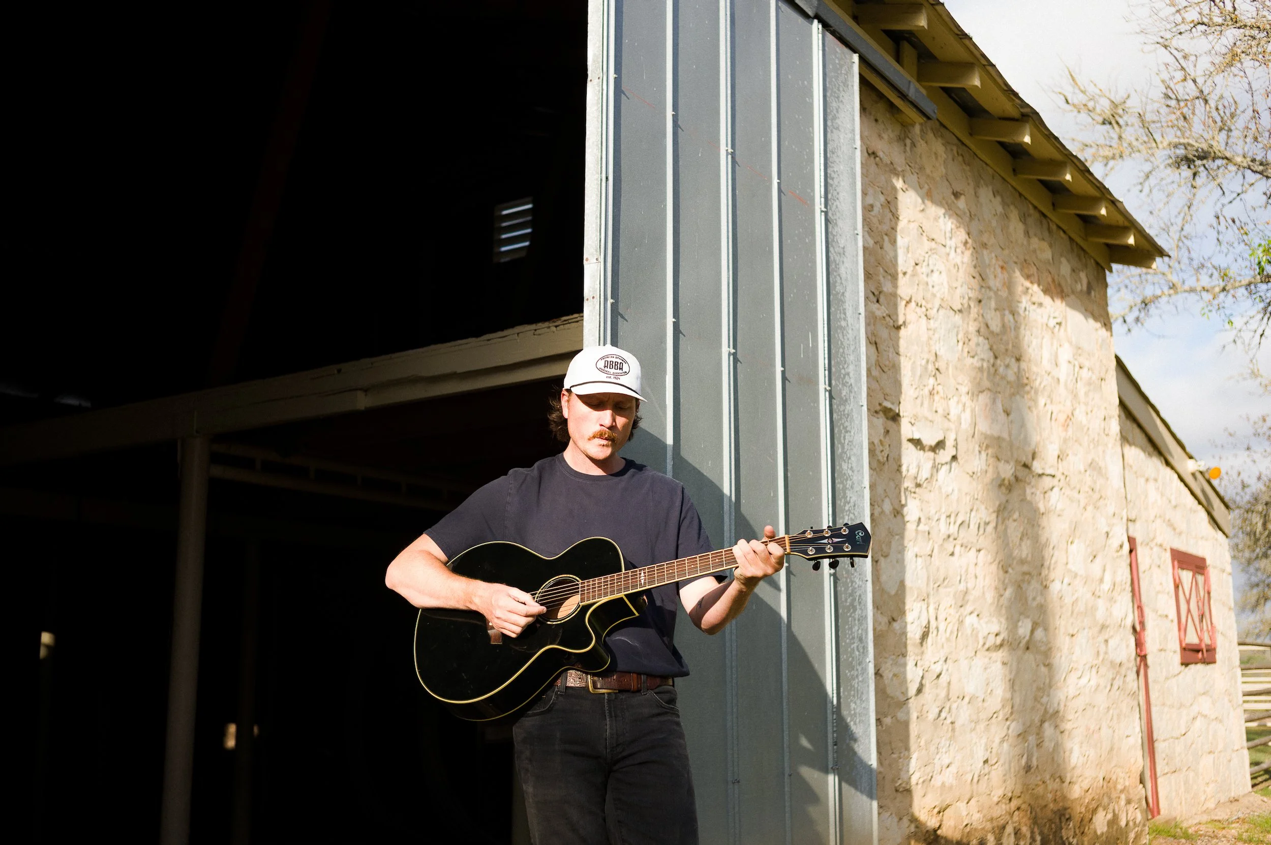 A man playing an acoustic guitar outdoors in front of a stone house under a clear sky.