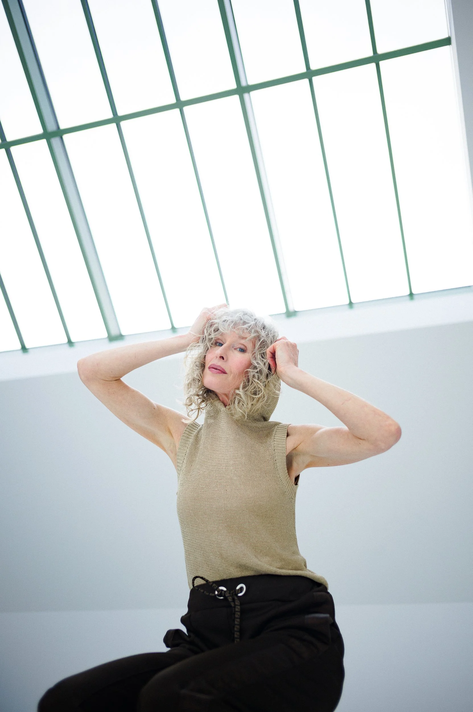 A woman with curly gray hair wears a beige sleeveless top and black pants, sitting cross-legged with hands behind her head, beneath a large skylight with a green metal grid.