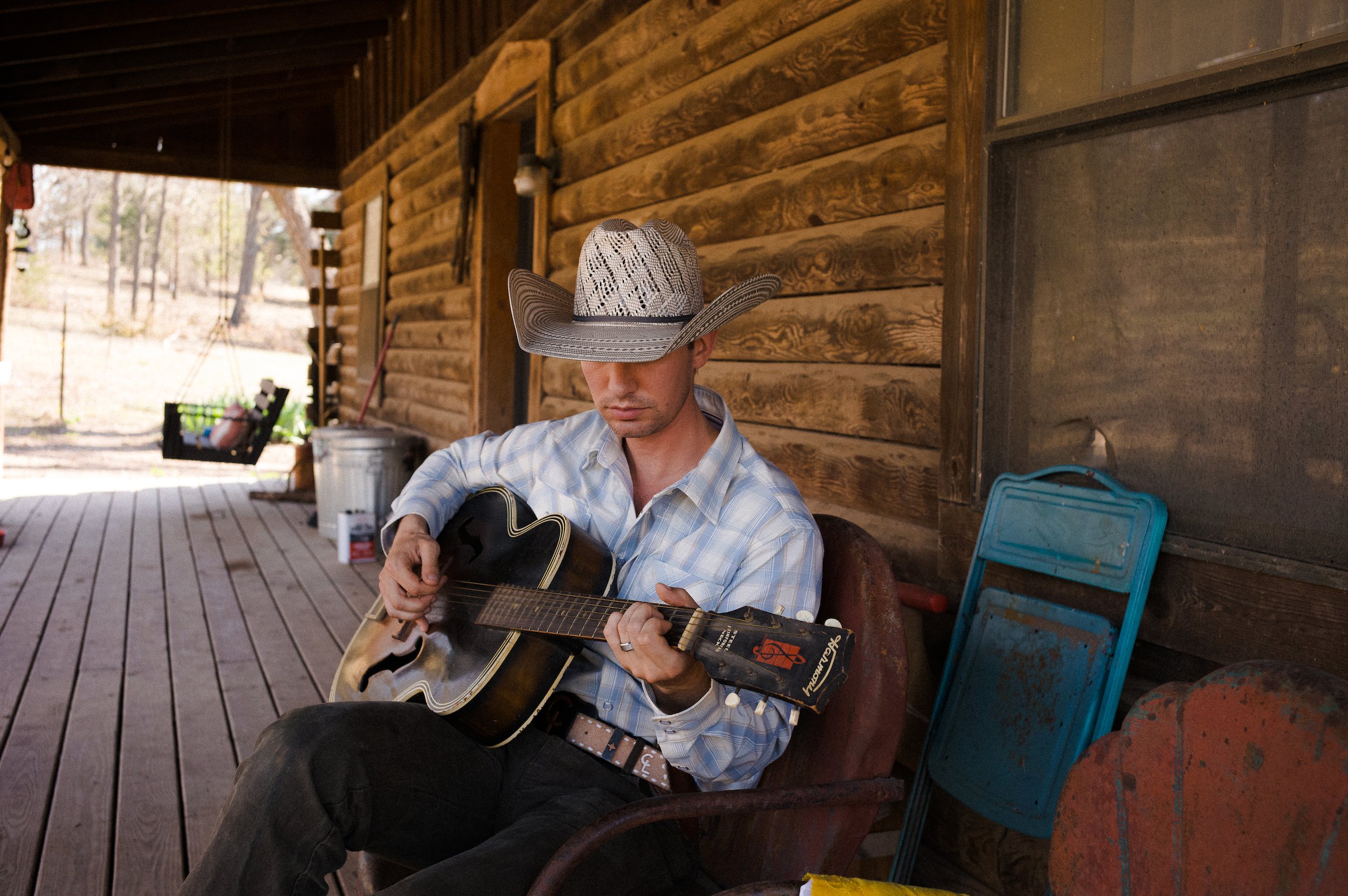 Man wearing a cowboy hat sitting on a porch, playing an acoustic guitar with wooden walls and outdoor scenery in the background.