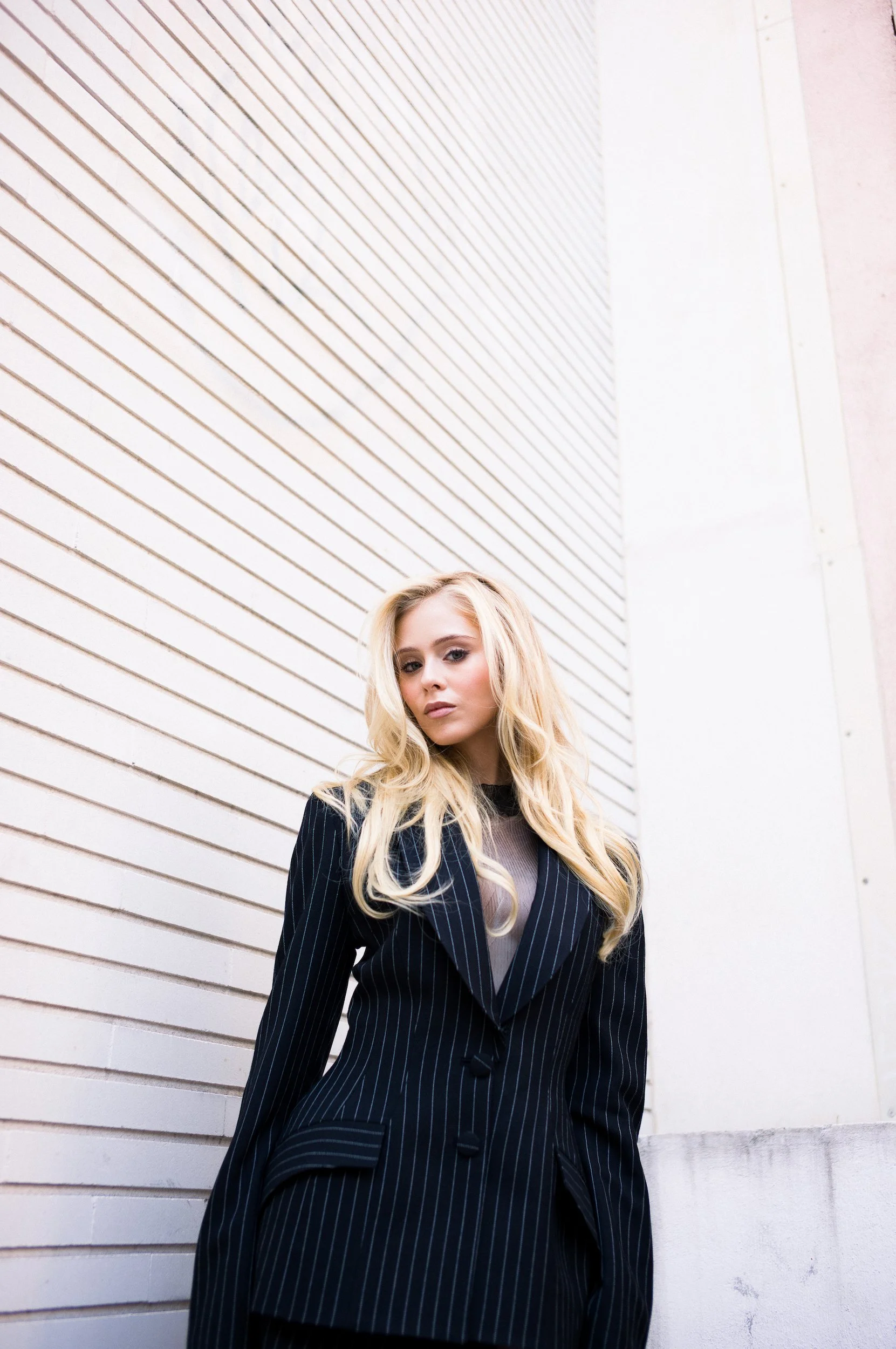 At Paris Fashion Week, A young woman with long blonde hair standing against a white wooden wall with diagonal lines, wearing a dark pinstripe blazer over a sheer top.