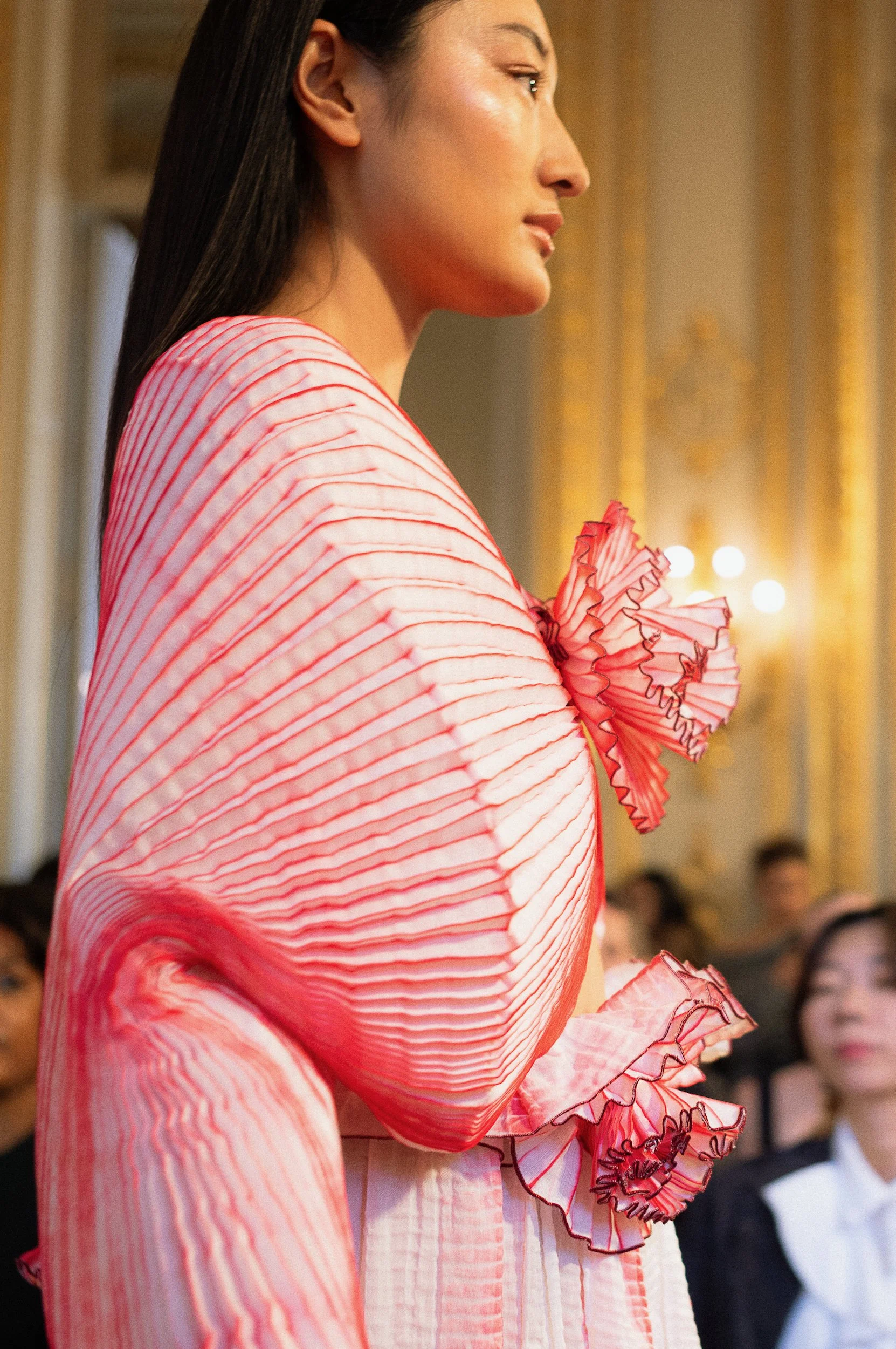 At Paris Fashion Week, Close-up side view of a woman wearing a pink and white striped dress with ruffled details, in a well-lit indoor setting, with an audience blurred in the background.