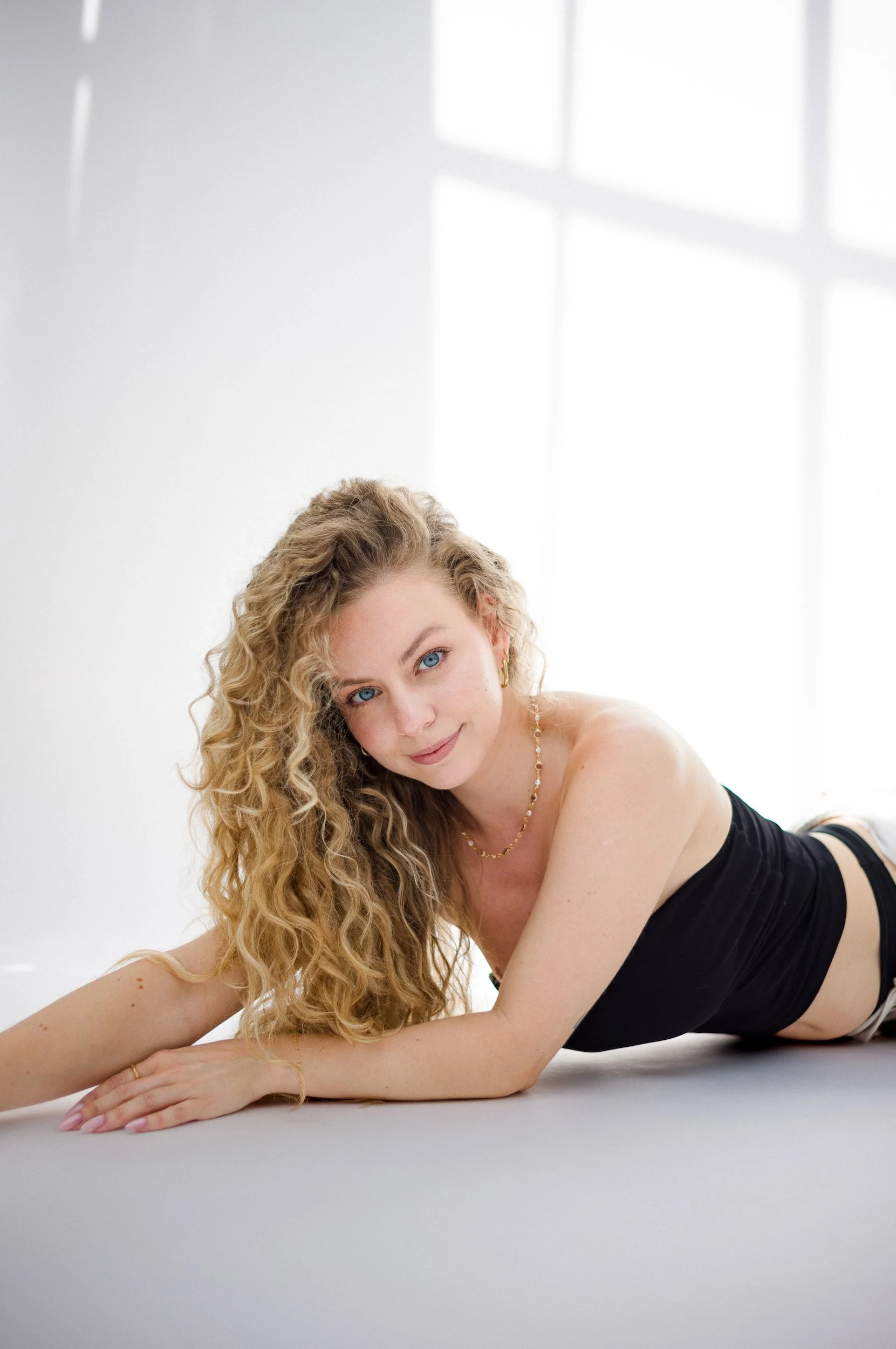 A woman with curly blonde hair and blue eyes lying on the floor, smiling gently, wearing a black strapless top, jewelry, and a ring, against a white background with natural light.