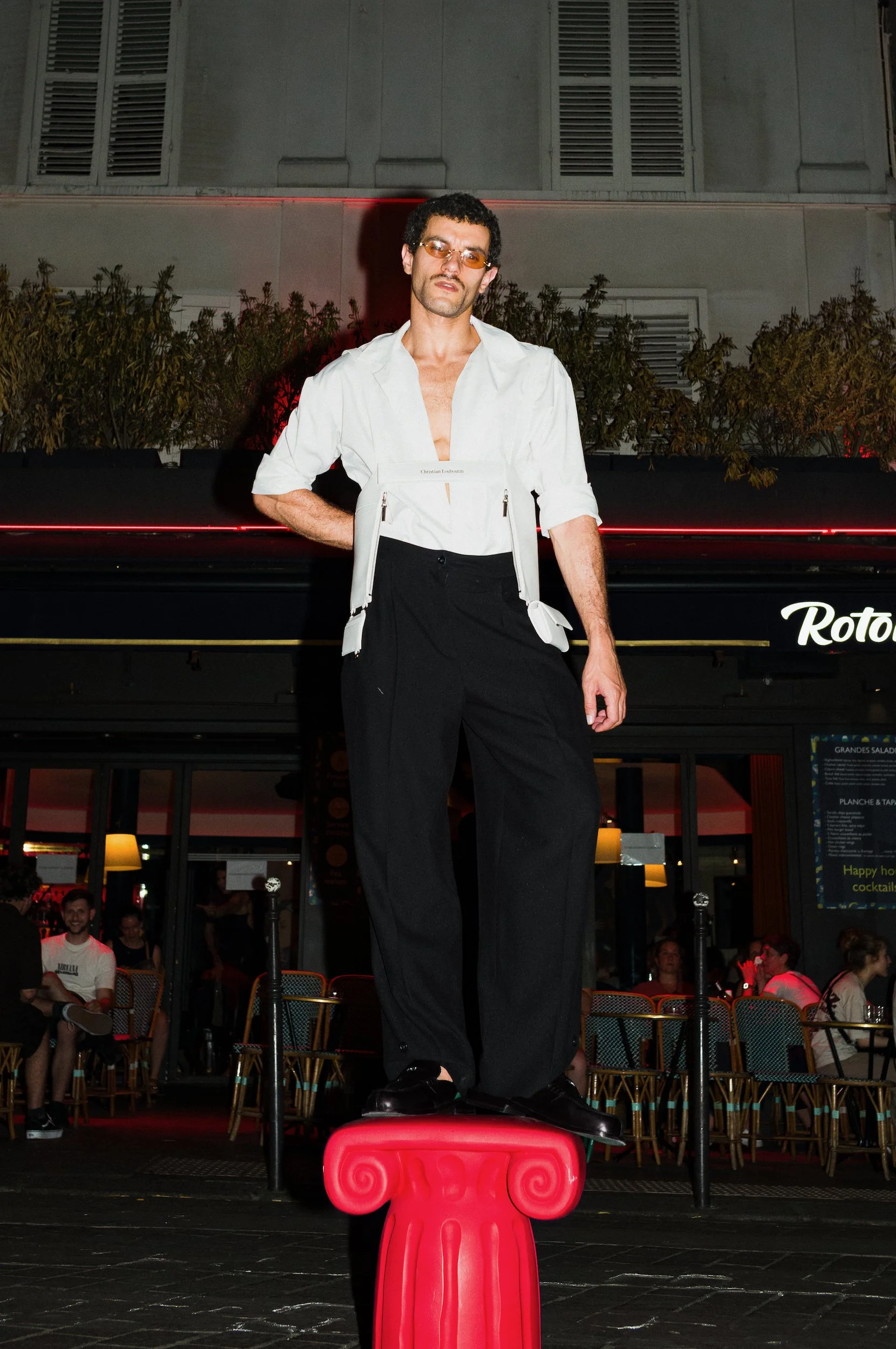 A man with glasses standing on a red pedestal with a pink spiral design, in front of an audience in a bar or restaurant setting at night.