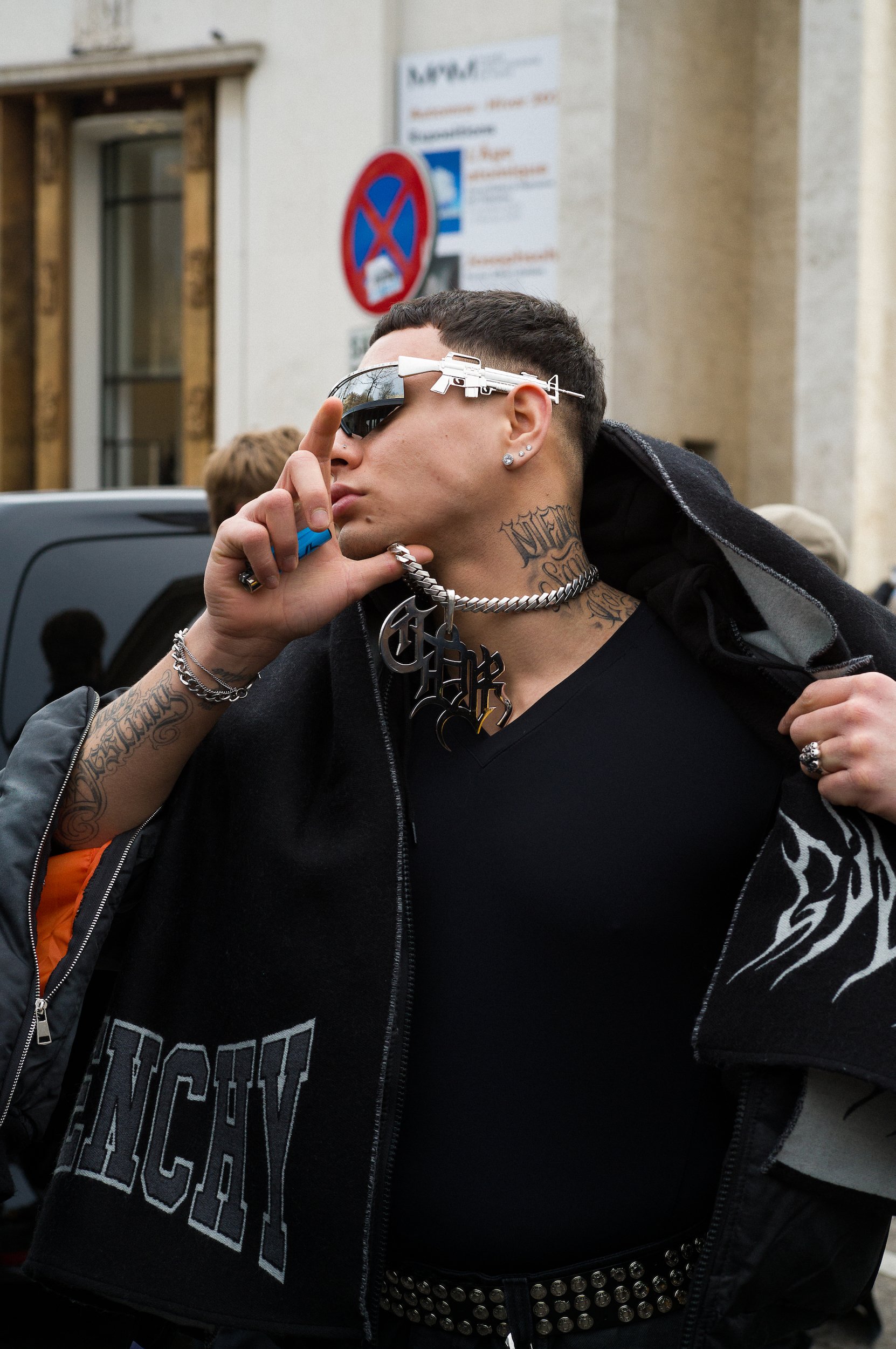 At Paris Fashion Week, Person with tattoos, studs, and a large chain wearing sunglasses with a gun-shaped reflection, standing outdoors in front of a building with signs.