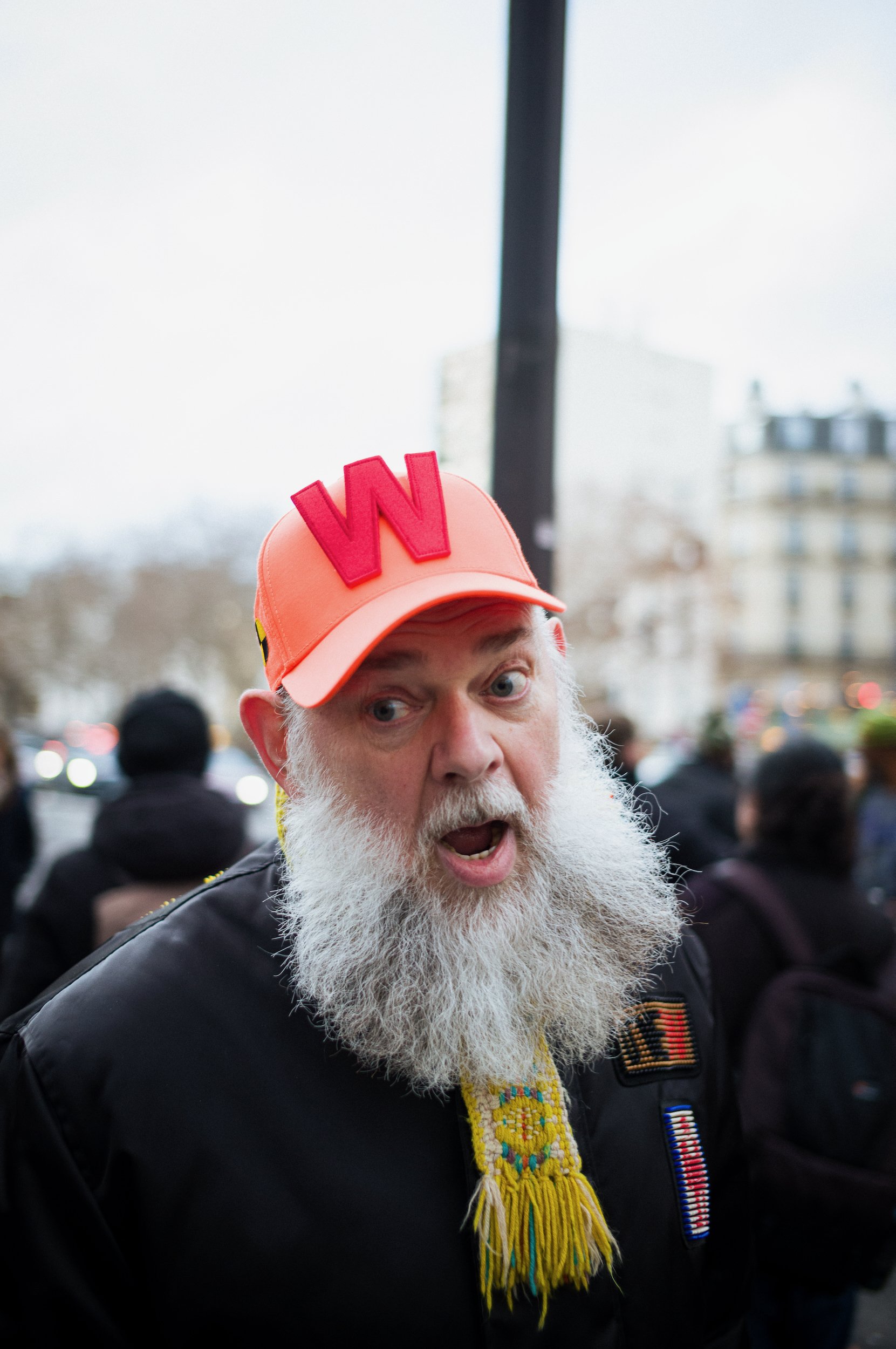 Walter Van Beirendonck, white beard, wearing an orange cap with a red 'W' on it, a beaded necklace, and a black jacket standing outdoors during daytime with a crowd and buildings in the background.
