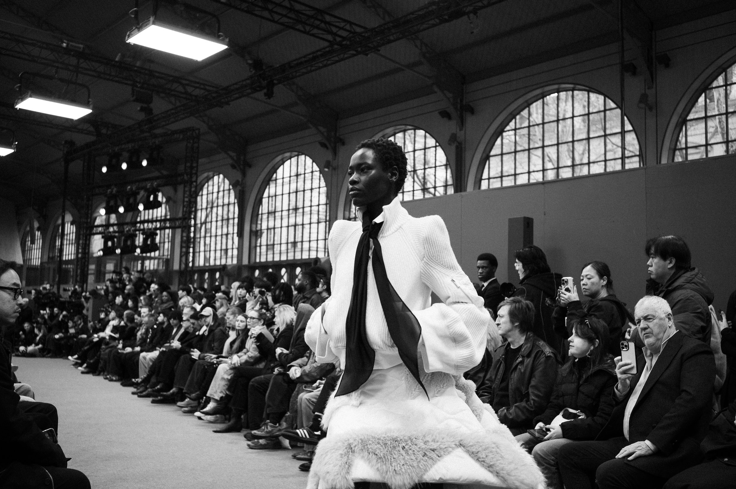 At Sacai for Paris Fashion Week, A woman walking on a runway during a fashion show in a large indoor space with arched windows and high ceilings, surrounded by seated spectators.