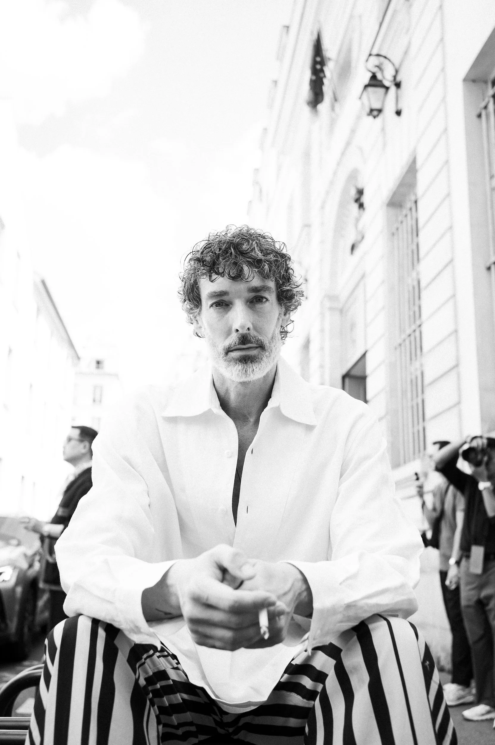 At Paris Fashion Week, A man with curly hair and a beard sitting cross-legged on the ground wearing a striped pair of pants and a white shirt, in an urban outdoor setting with other people around.