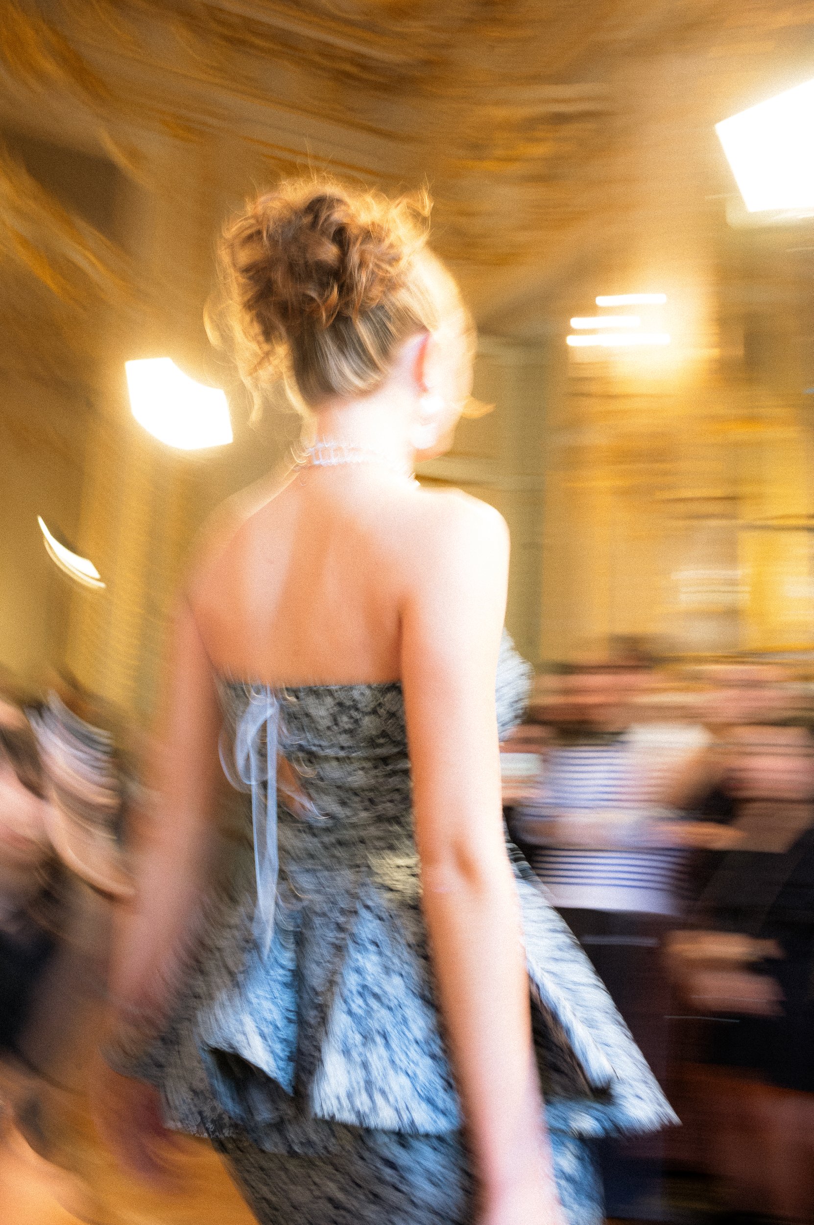 At Paris Fashion Week, A woman with curly hair tied up in a bun, wearing a strapless top and a choker necklace, sitting in a room with warm-colored lighting and blurred background.